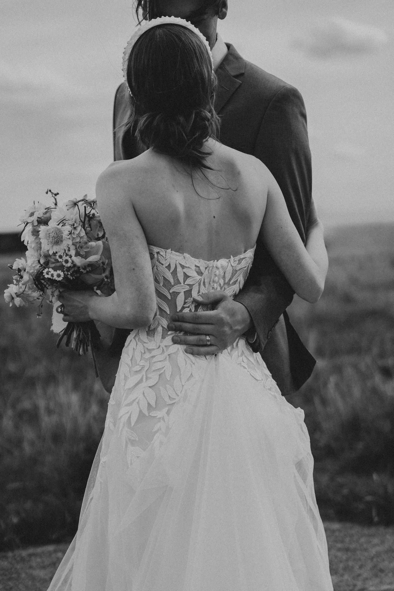 Black and white photo of bride in lace strapless gown holding bouquet embraced by groom in field.