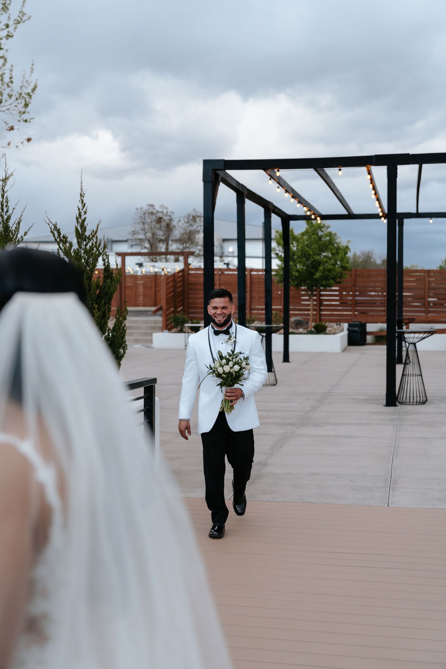 Smiling groom in white tuxedo jacket walks toward bride holding bouquet on outdoor wedding venue deck.