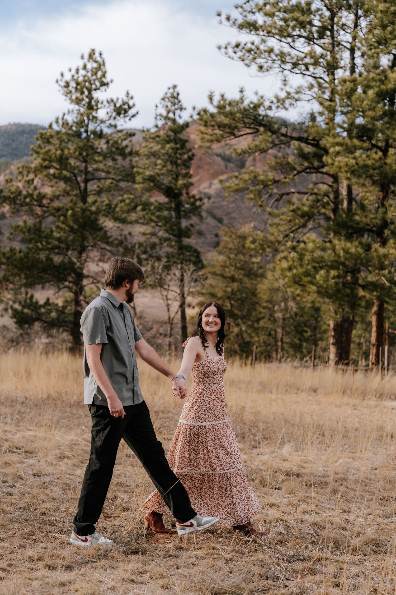 Smiling woman in floral maxi dress walking hand in hand with man through Colorado mountain meadow.