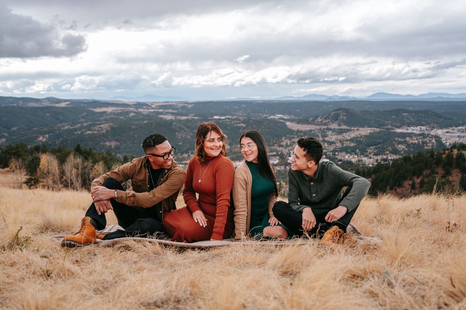 Four friends sitting on a hillside with panoramic mountain valley views on a cloudy day.