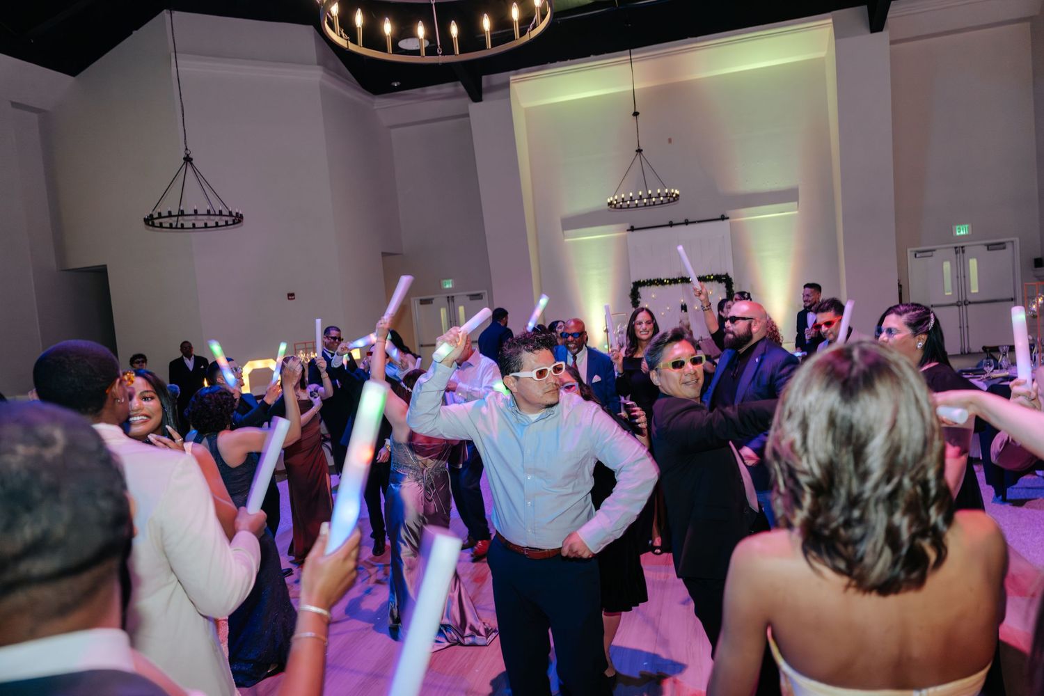Guests dancing and celebrating at a lively wedding reception with glow sticks and sunglasses indoors.