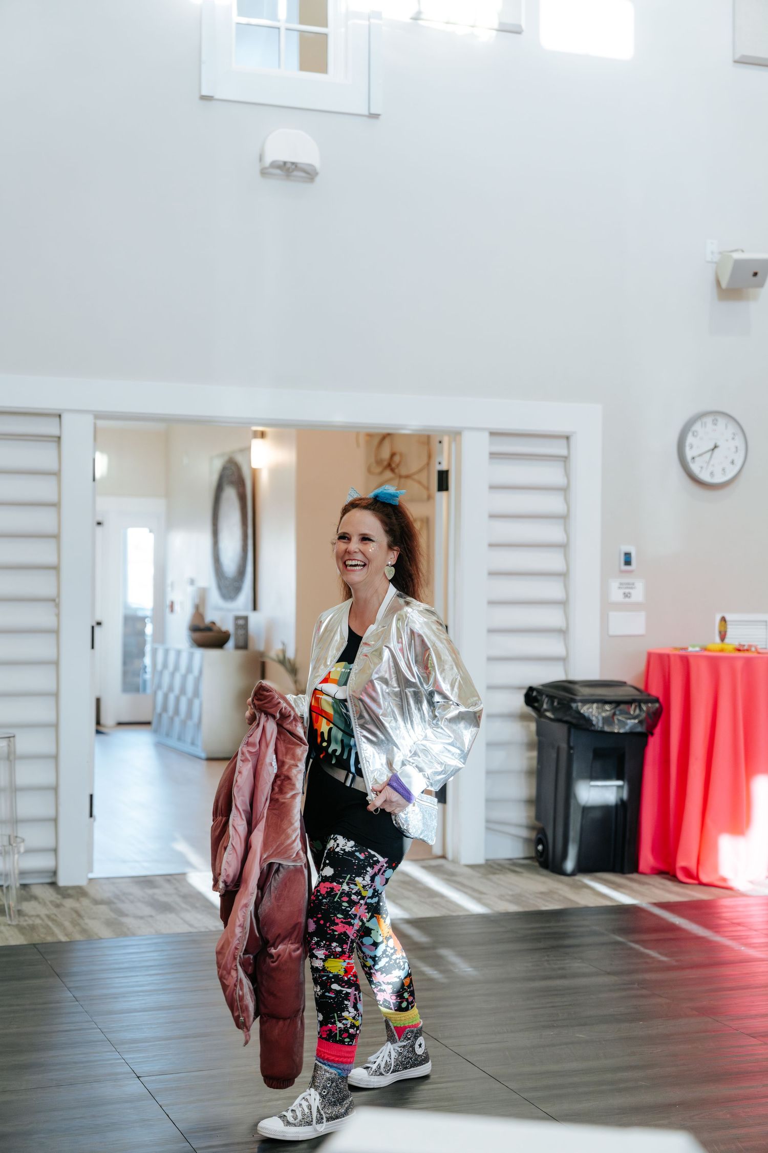 Woman in silver jacket and floral pants smiling in bright modern venue interior.