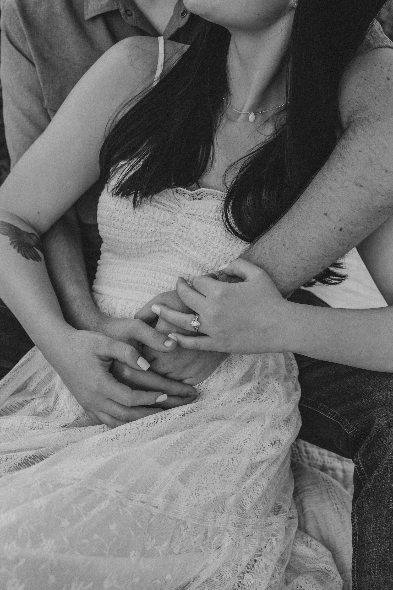 Black and white photo of a couple holding hands, woman in white lace dress with man embracing her from behind.