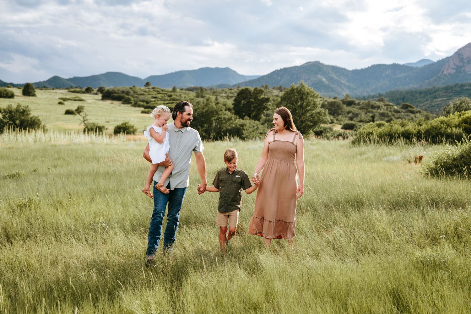 Happy family of four walks through lush green mountain meadow on a cloudy summer day.