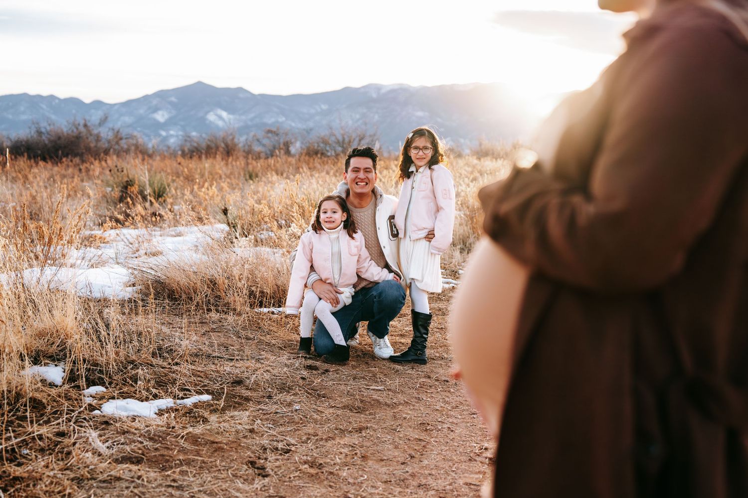 Father crouching with two young girls smiling outdoors, mountains and golden grass in the background at sunset.