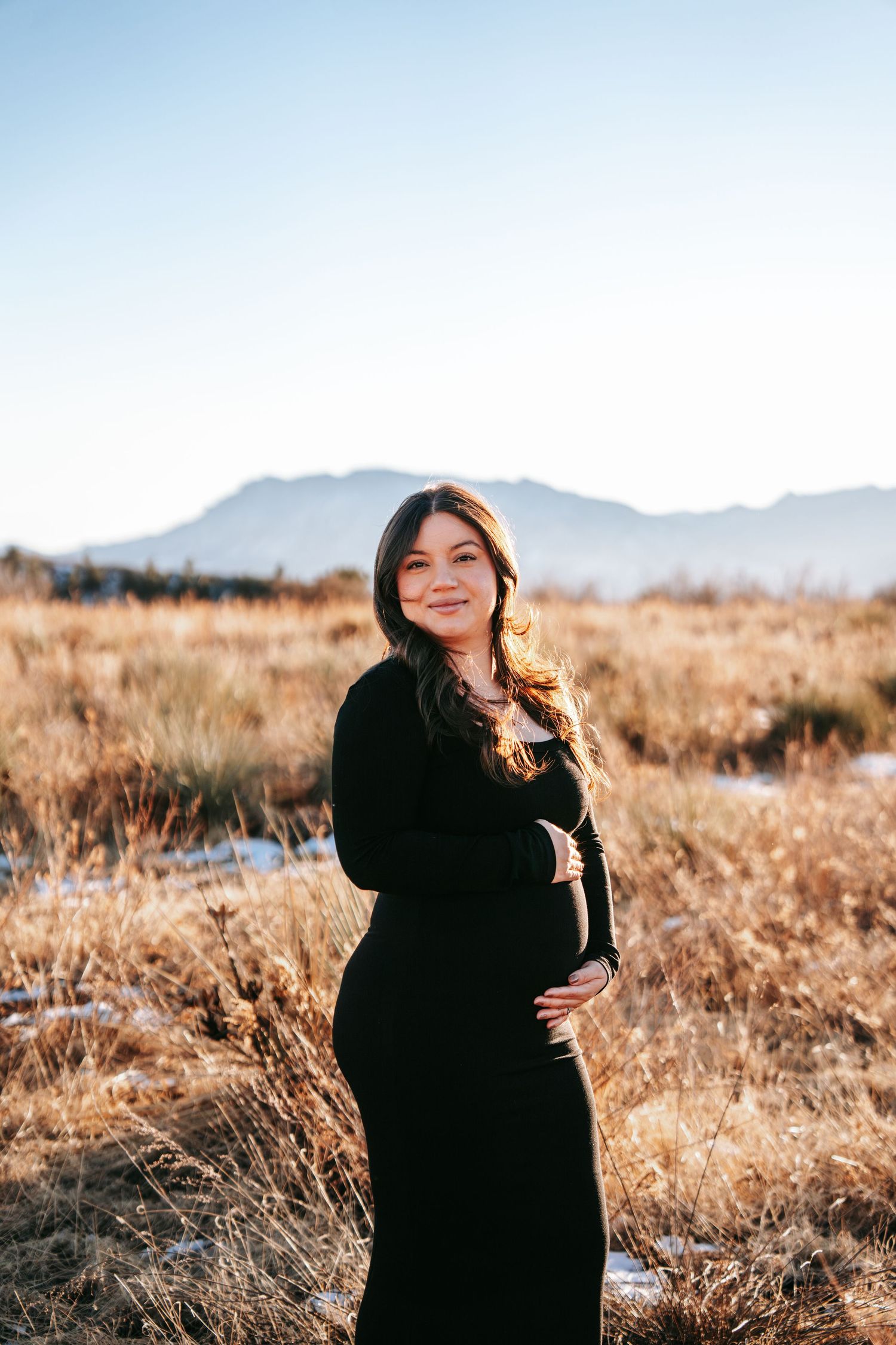Pregnant woman in black maxi dress cradling baby bump in sunlit winter field with mountains.