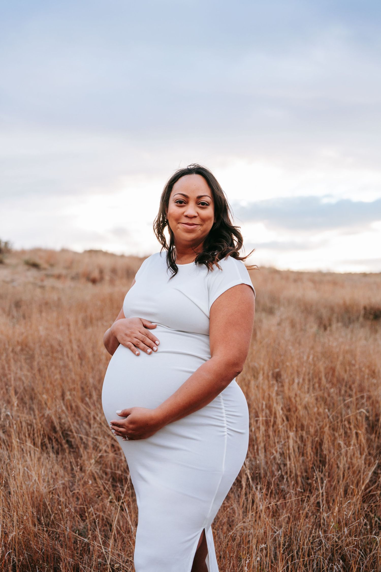 Pregnant woman in white fitted dress holds baby bump while standing in golden dry grass field.
