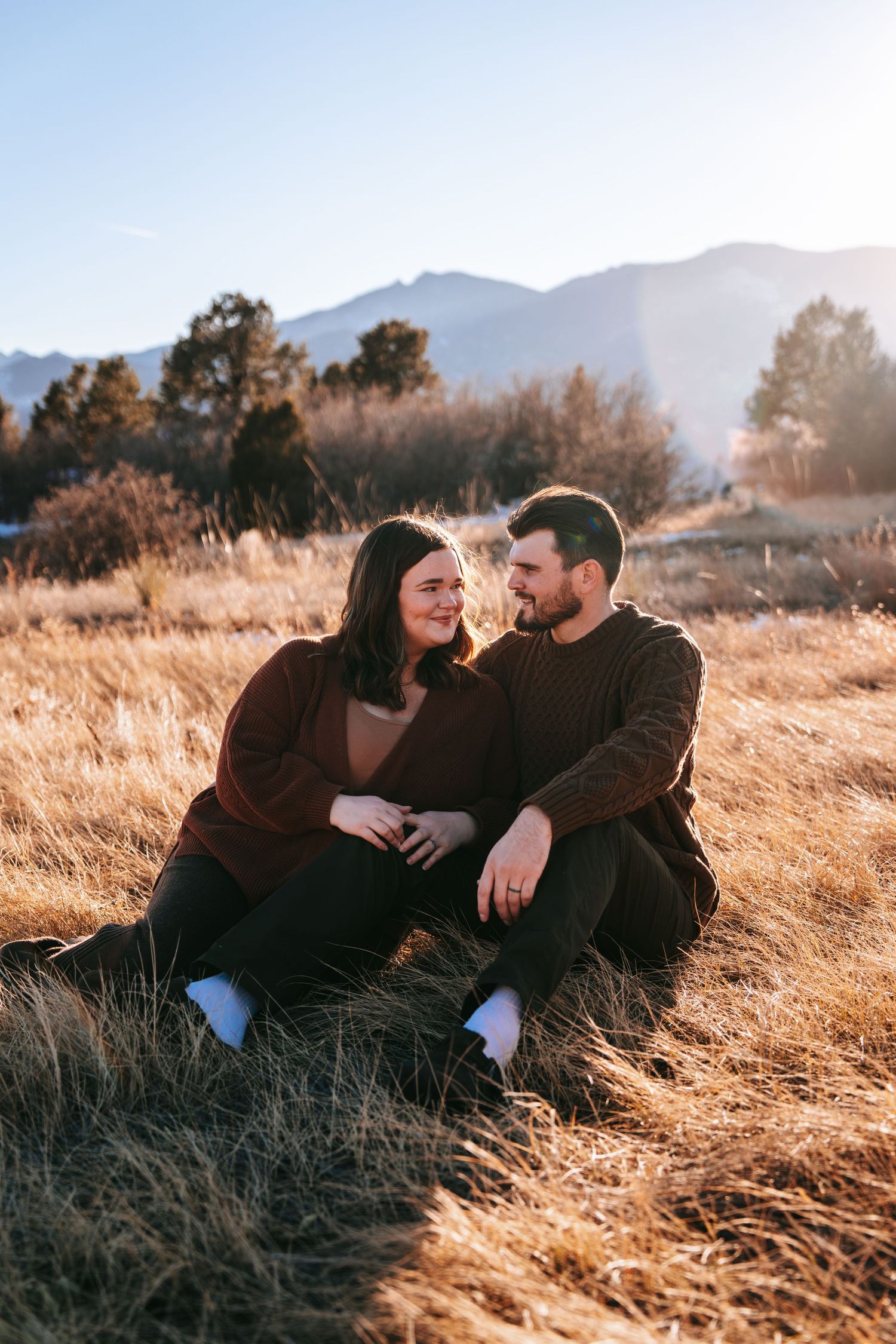 Couple sitting together in golden grass field with mountain backdrop in winter sun.