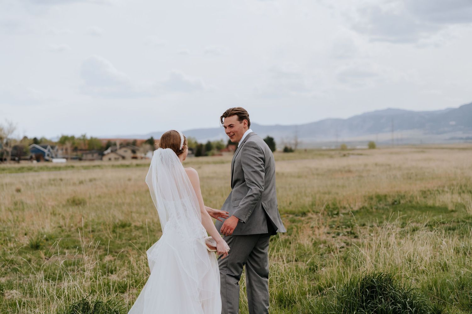 Bride in flowing white veil and groom in gray suit hold hands while walking through a scenic meadow.