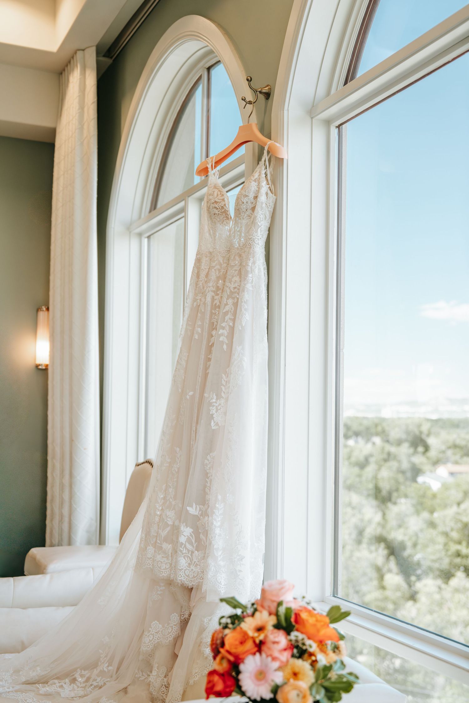 Lace wedding dress hanging on wooden hanger in arched window with blue sky and colorful bouquet below.