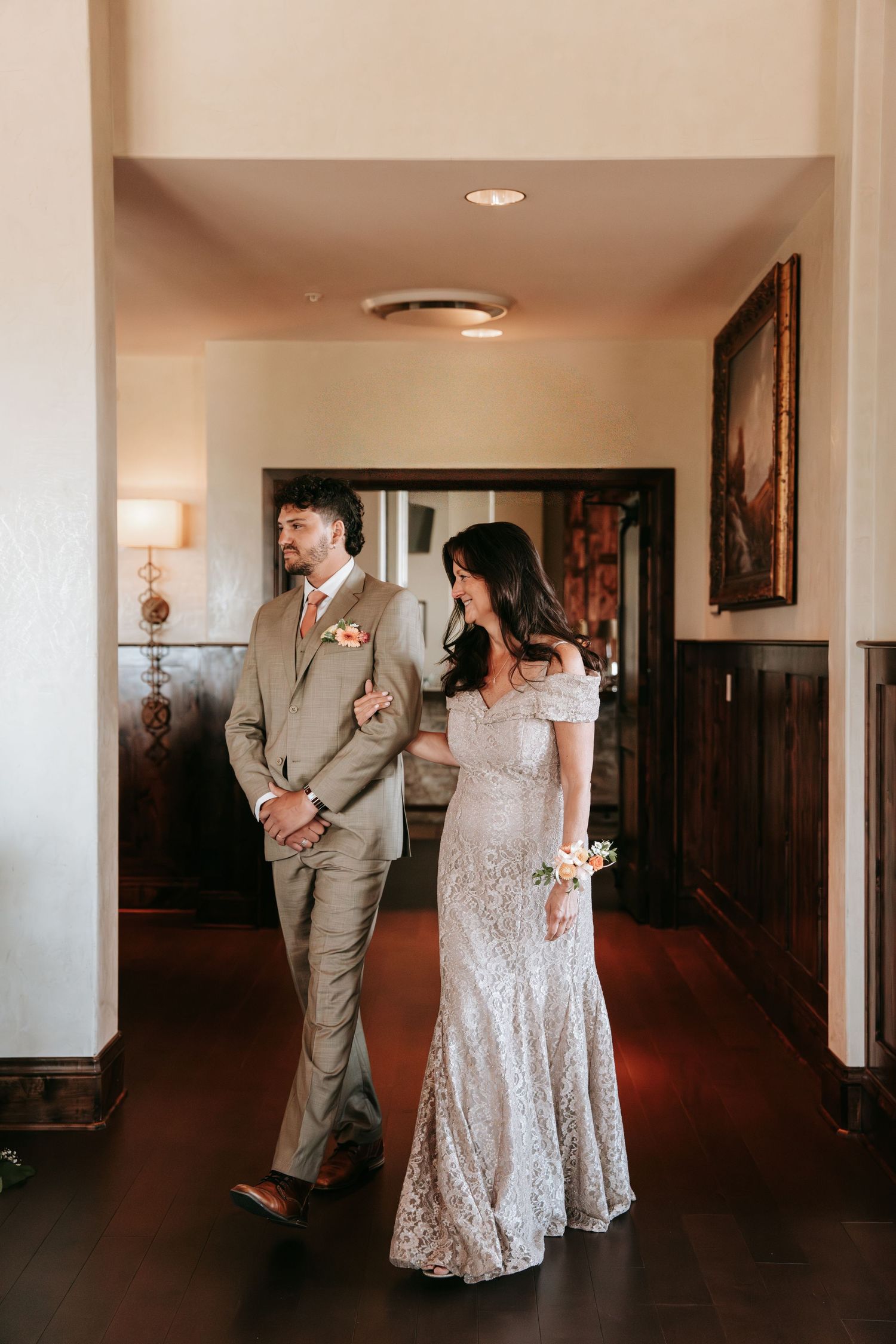 Bride in off-shoulder lace gown walks with groomsman in tan suit through a warmly lit hotel corridor.