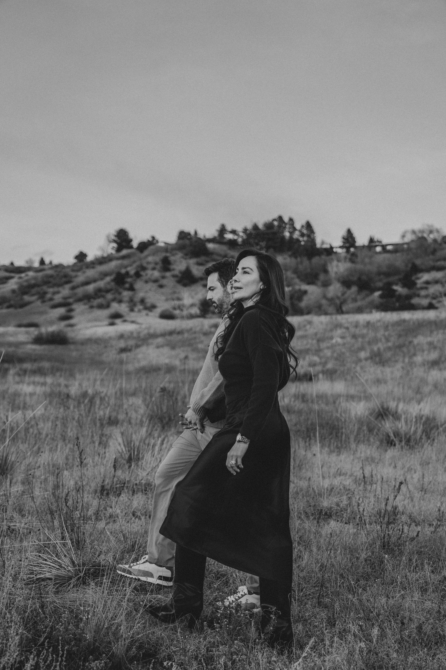 Black and white romantic photo of couple embracing in open grassy field with rolling hills behind them.