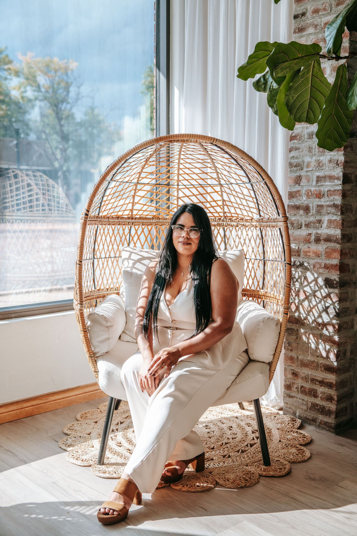 Woman in white outfit sitting in a wicker egg chair near a window with brick wall and plant.