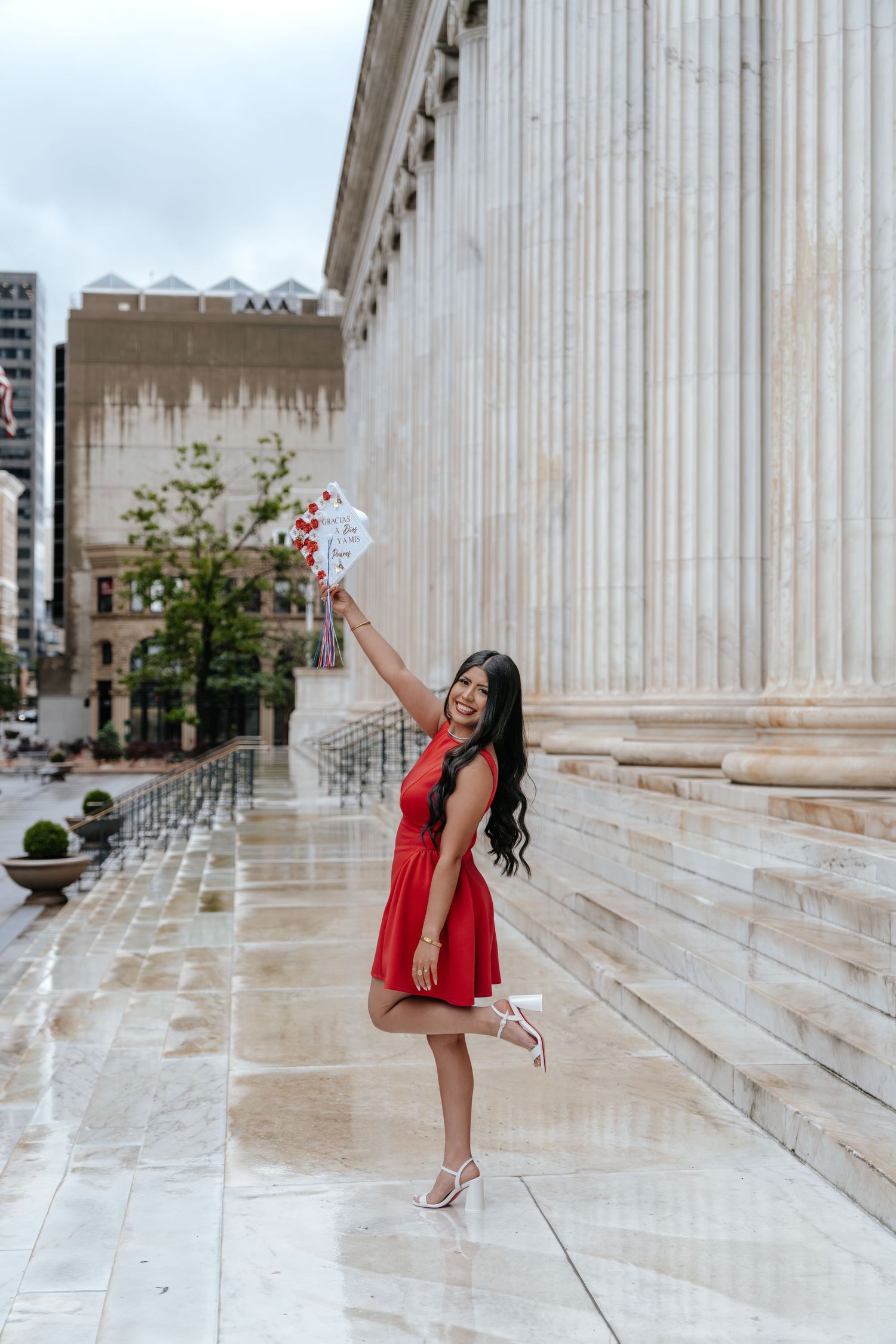Joyful woman in red dress raising graduation cap outside neoclassical building on rainy day.