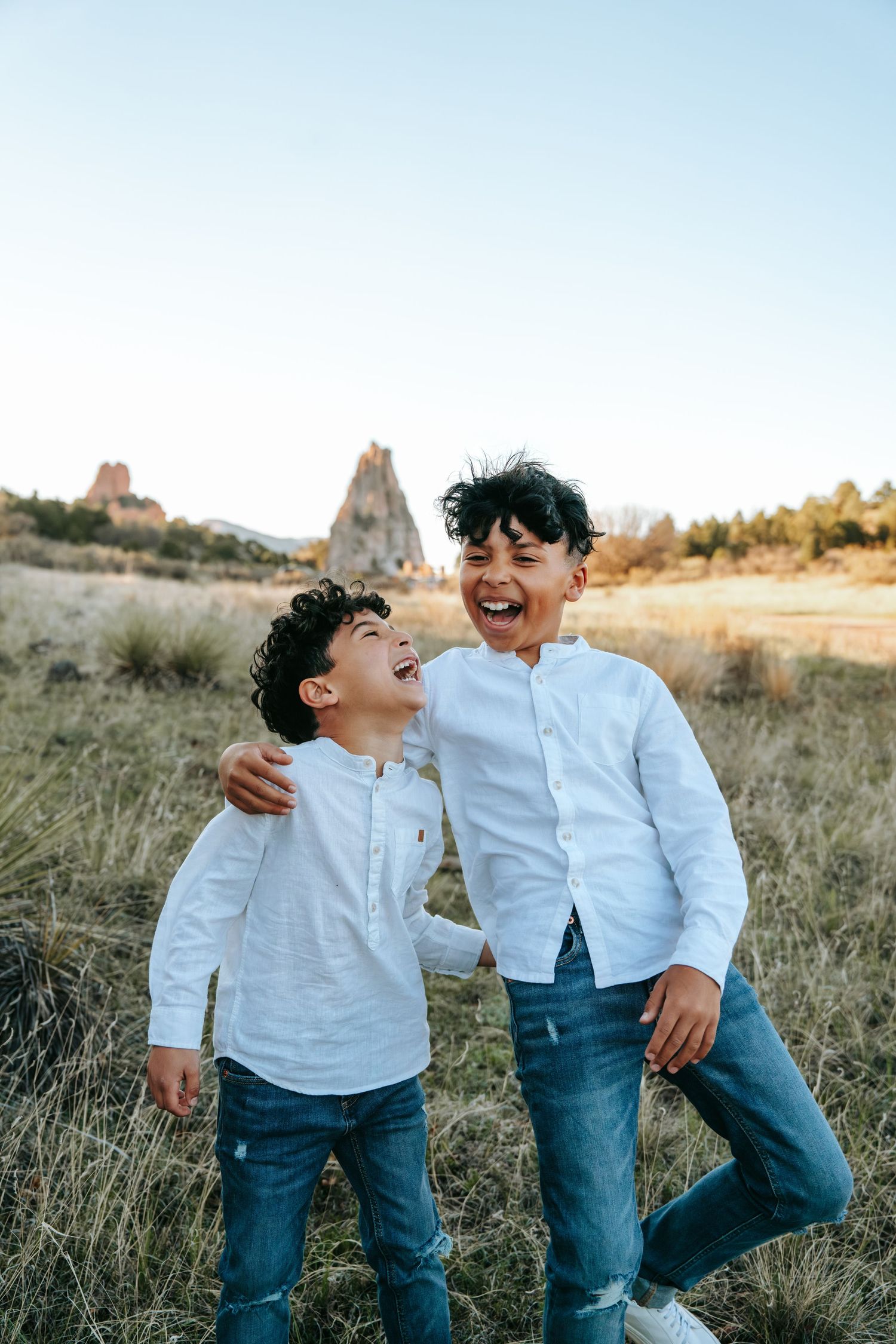 Two laughing boys in white shirts and jeans embrace outdoors with rocky landscape behind them.