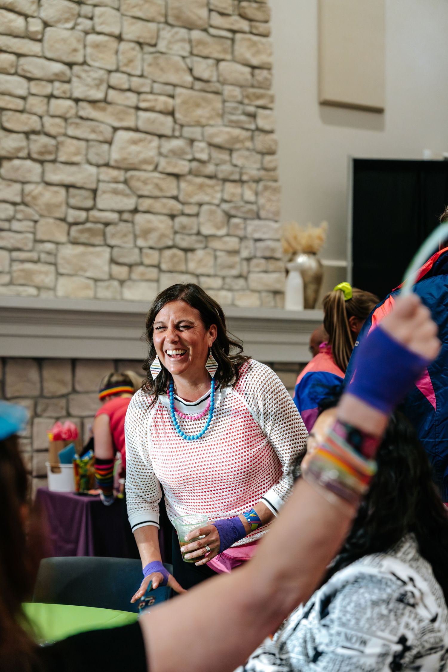 Woman in colorful 80s-themed outfit laughing at a retro party near a stone fireplace.