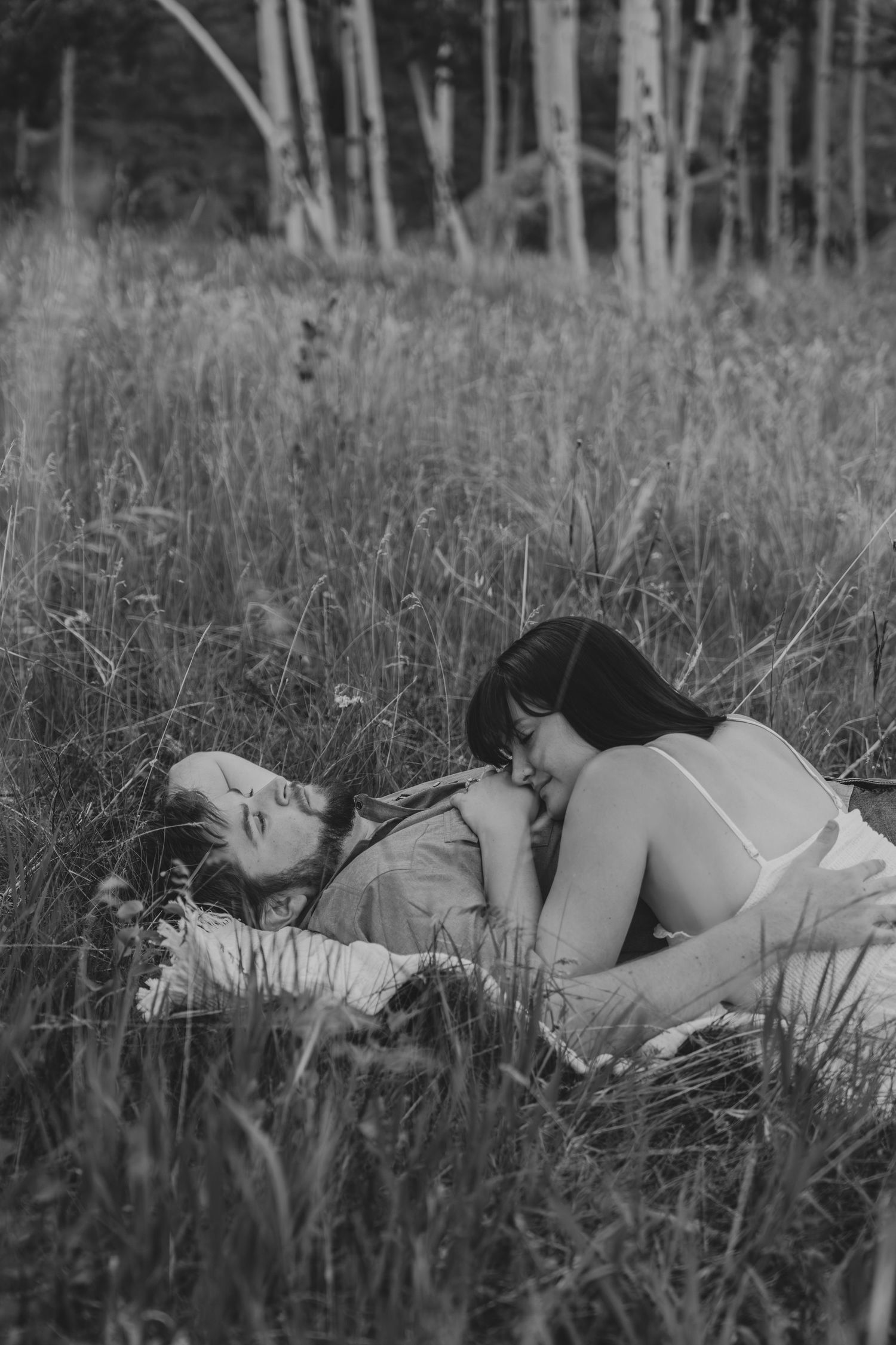 Black and white photo of two women lying in a grassy meadow with birch trees, sharing an intimate moment.