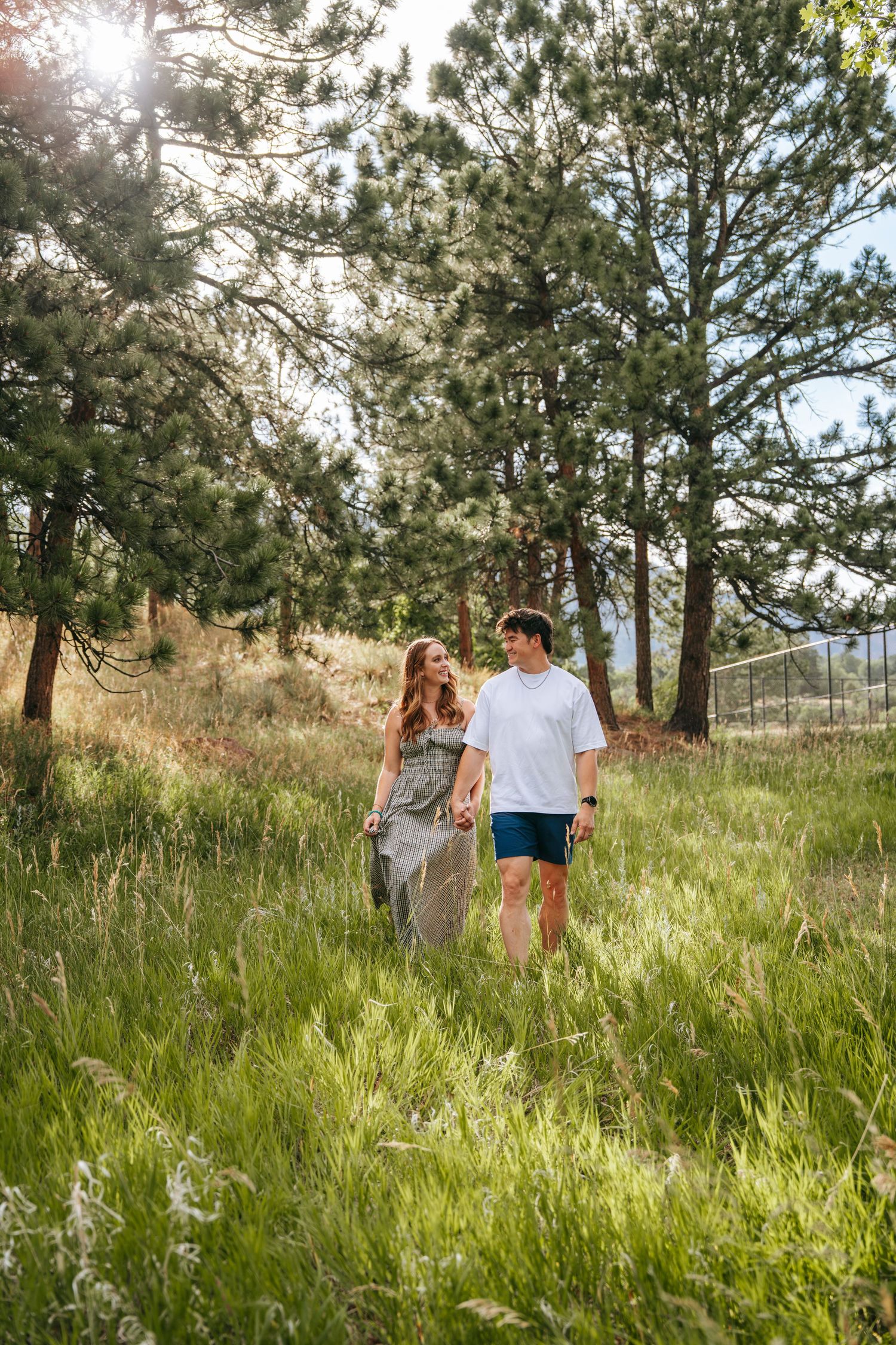 A couple walks hand-in-hand through a lush green meadow surrounded by tall pine trees in warm sunlight.