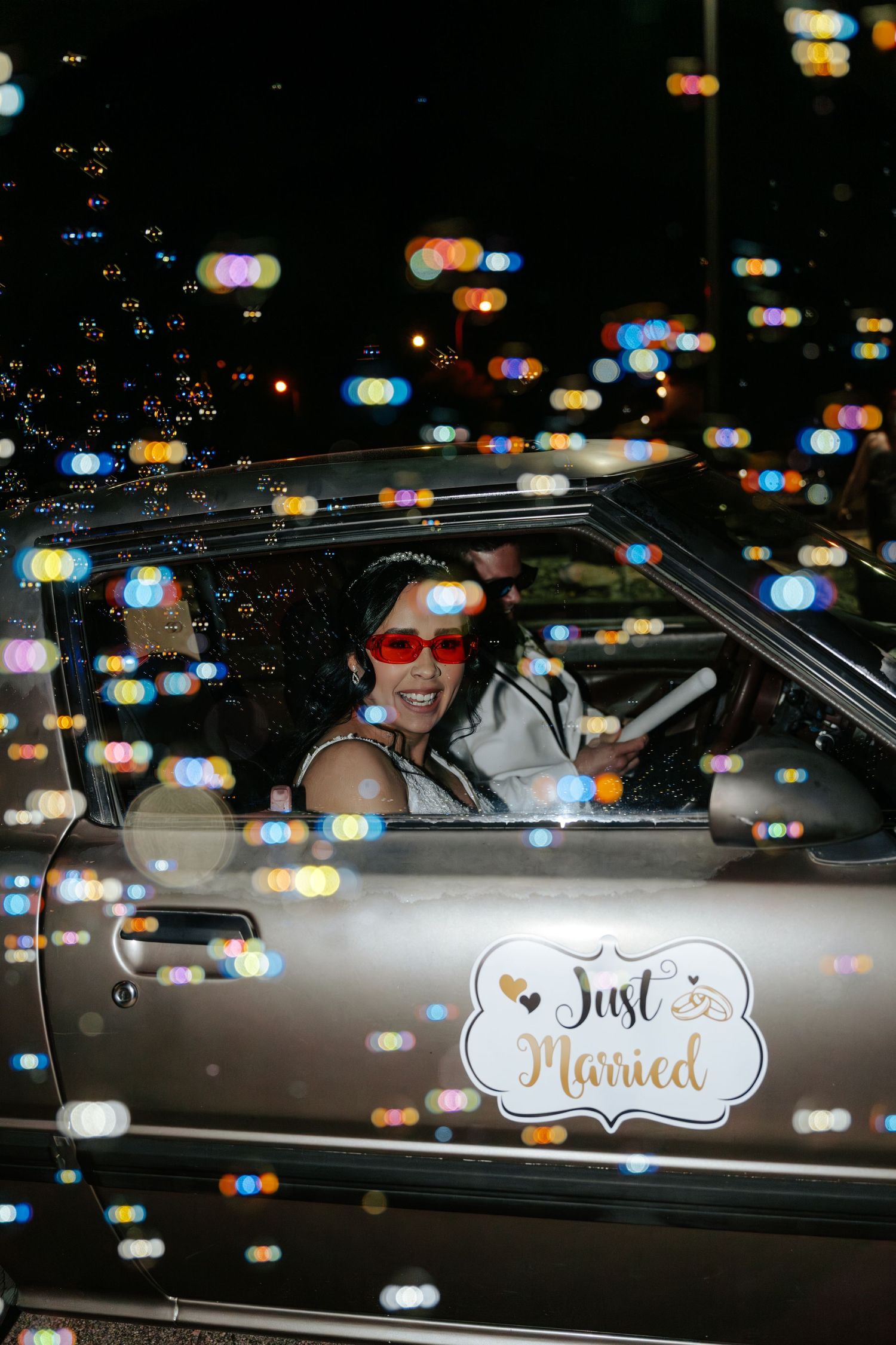 Smiling bride in a Just Married car departs wedding reception at night surrounded by colorful bokeh lights.
