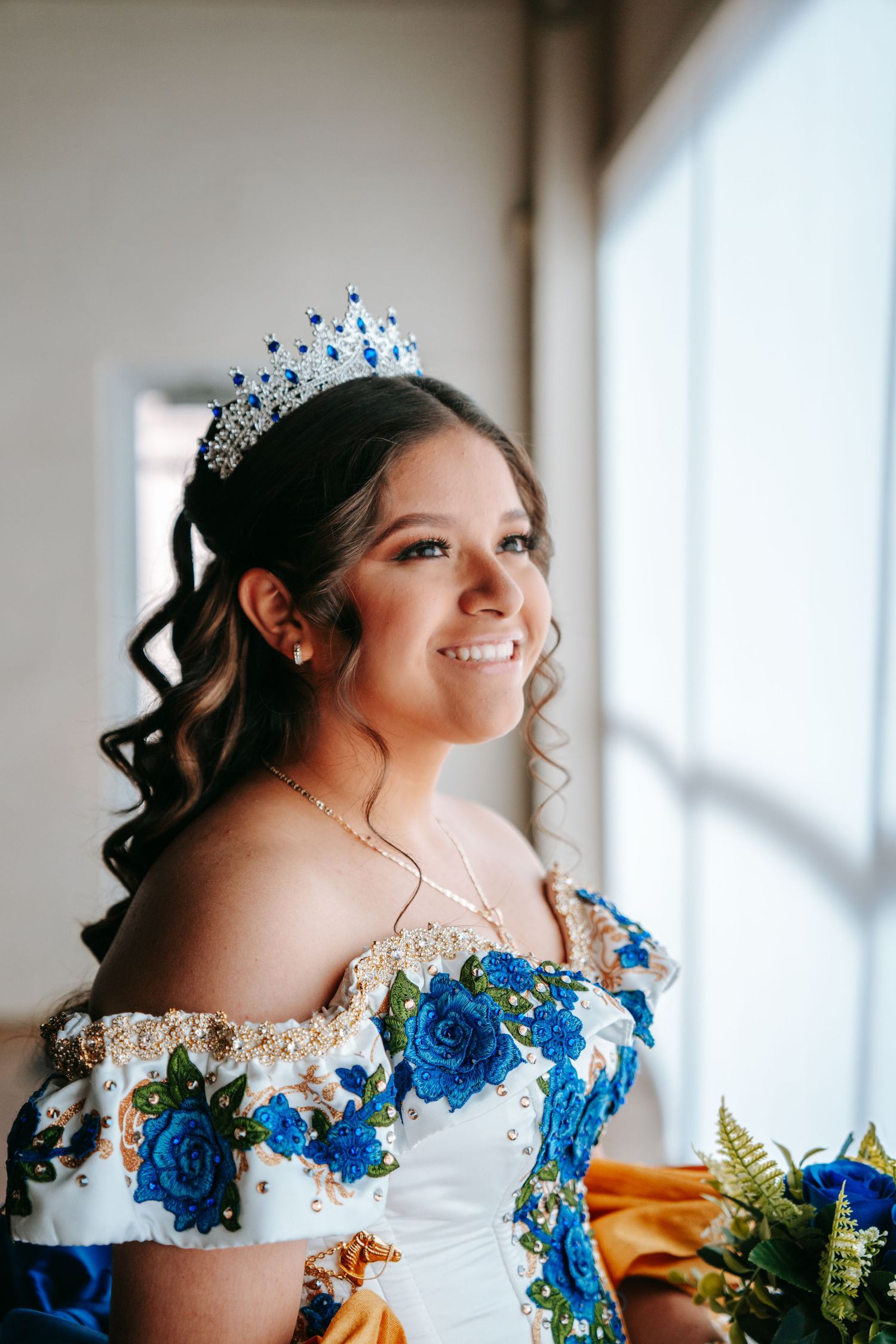 Young woman in blue floral quinceañera gown and tiara smiling by a bright window.