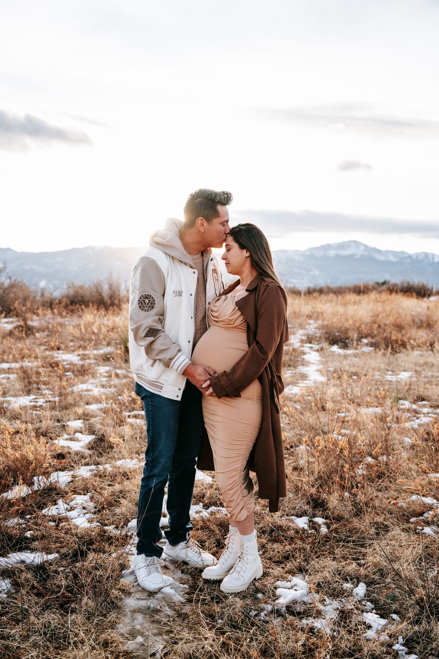 Couple sharing a tender moment in snowy field at sunset, man kissing pregnant womans forehead near mountains.