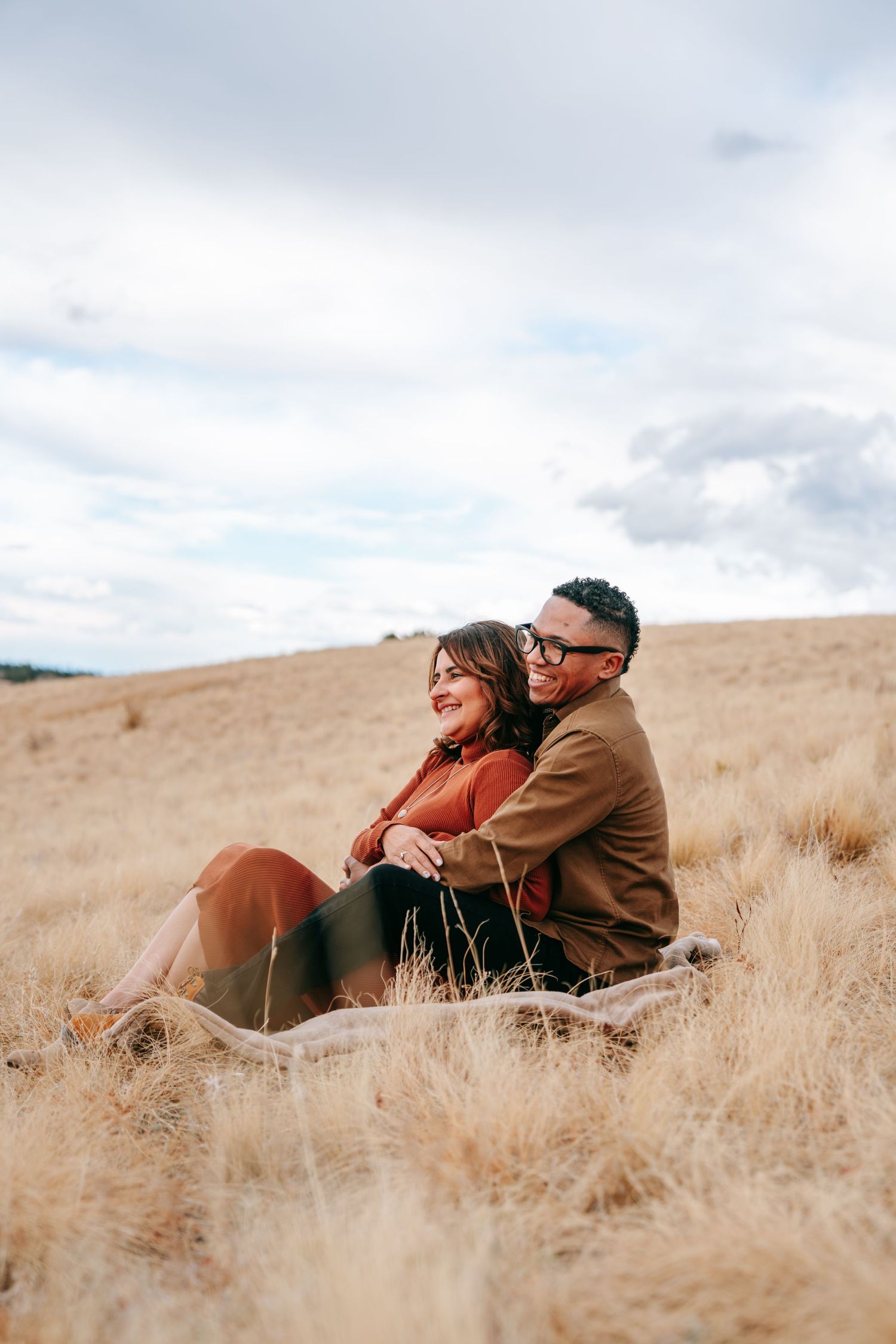 Couple sitting together in a golden grass field, laughing and cuddling with cloudy skies above.