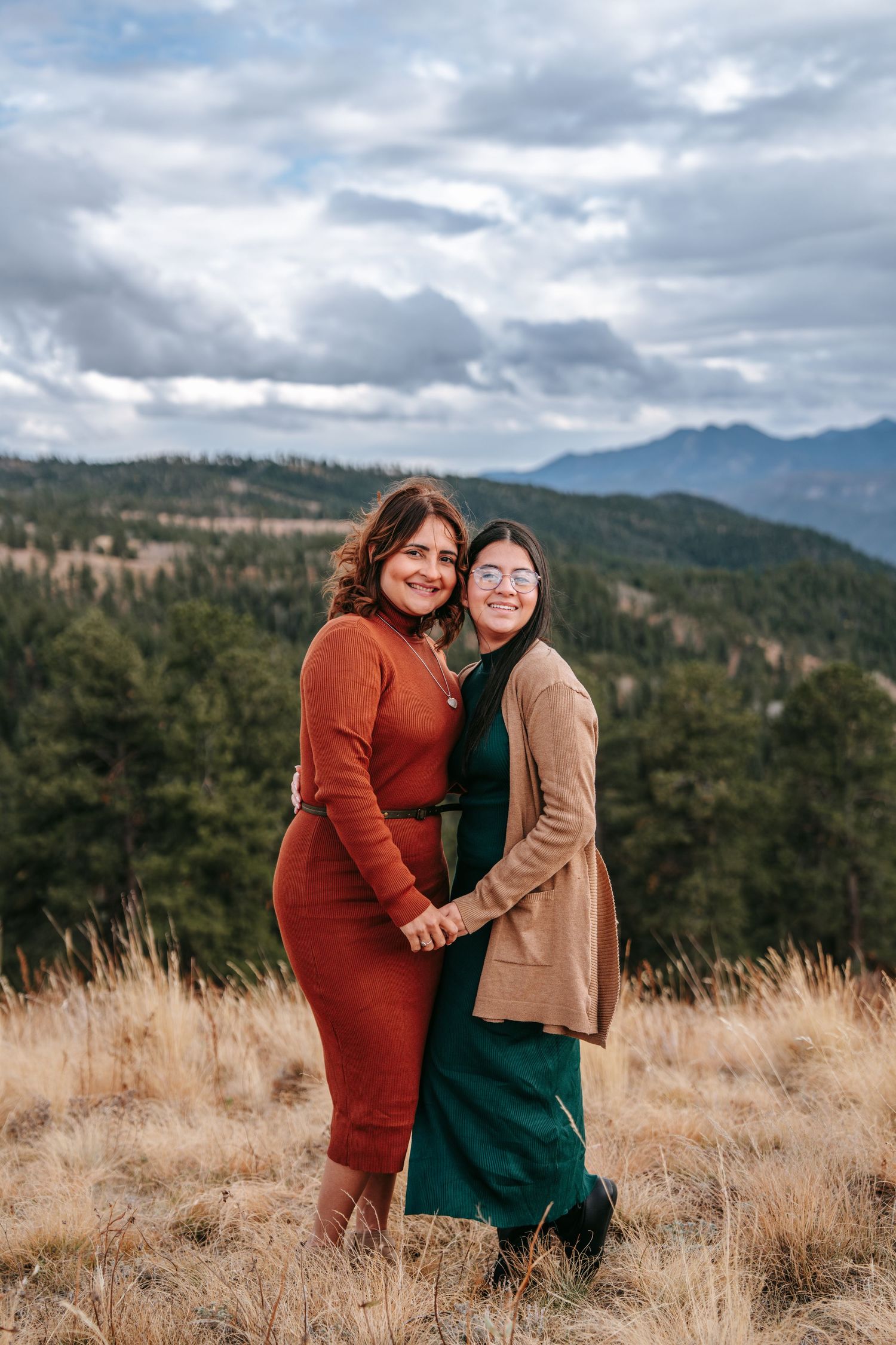 Two women posing together holding hands in a mountain meadow with forested hills in the background.