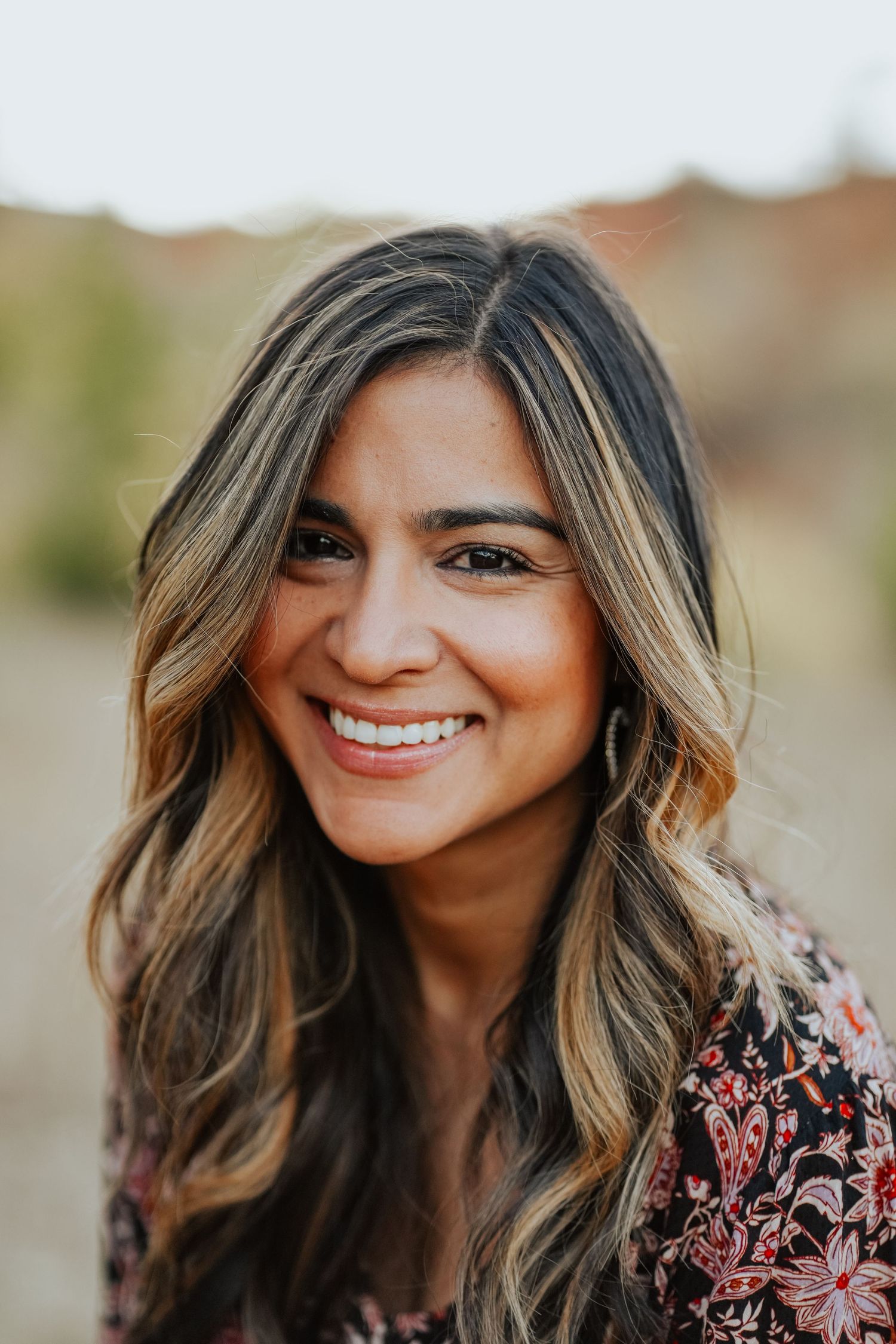 Close-up portrait of smiling woman with highlighted wavy hair in floral top outdoors.
