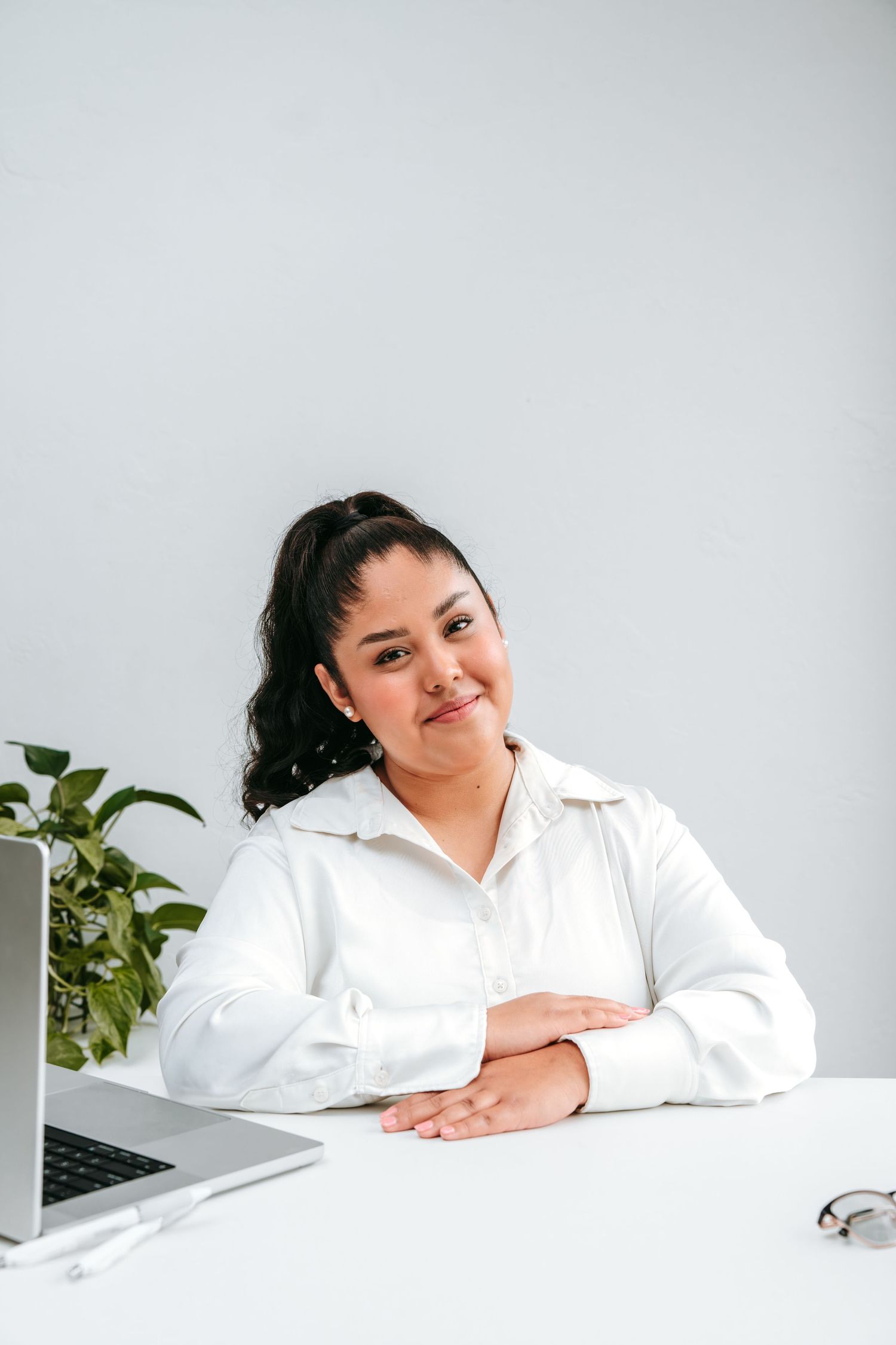 Professional woman in white shirt sitting at a white desk with a laptop and plant behind her.