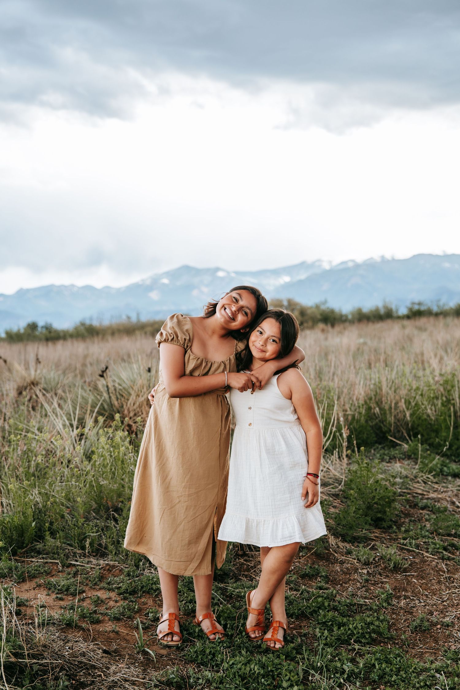 Two smiling girls hugging in a grassy field with snow-capped mountains visible in the background.