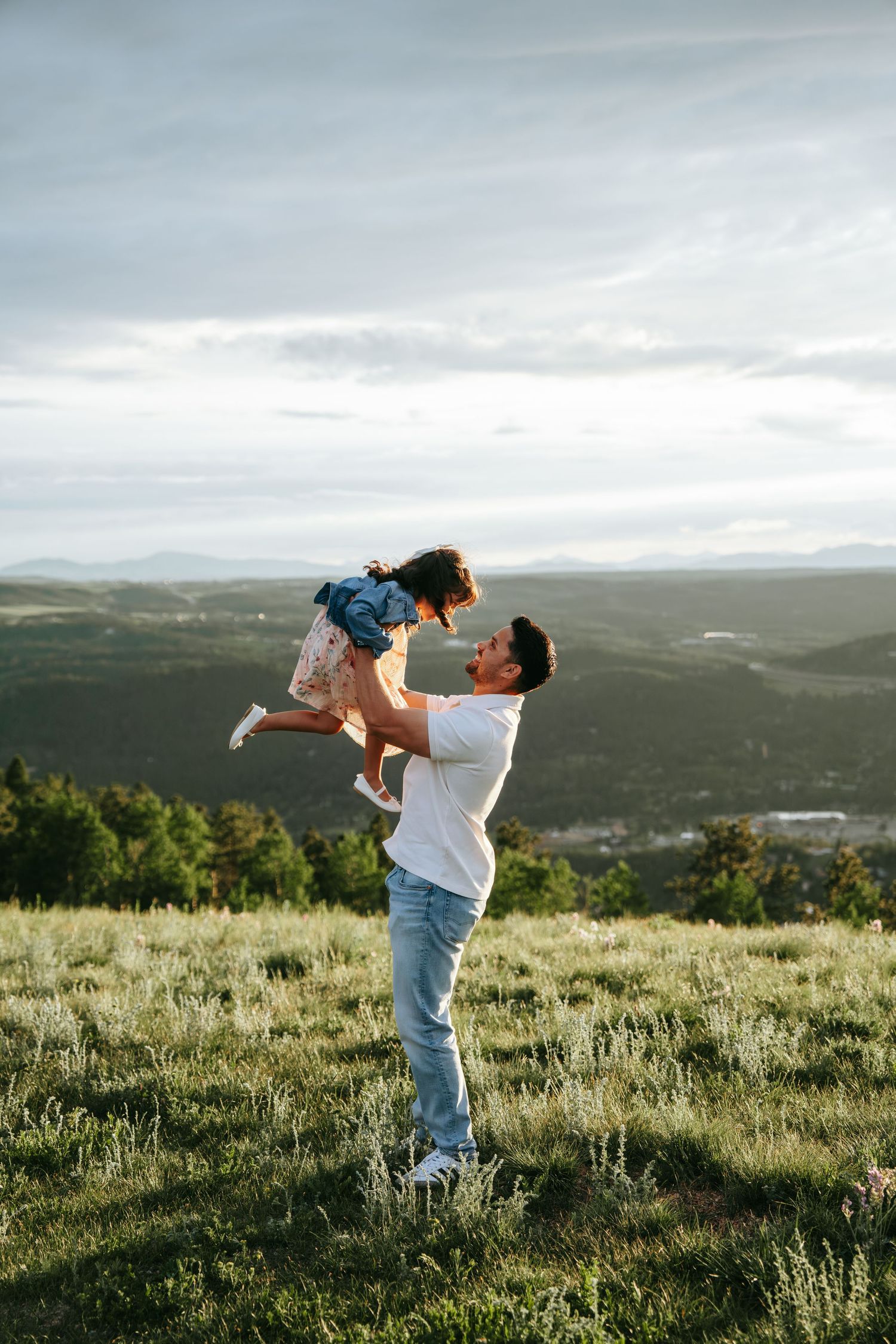 Father lifting young daughter into the air on a green hillside with panoramic mountain landscape.