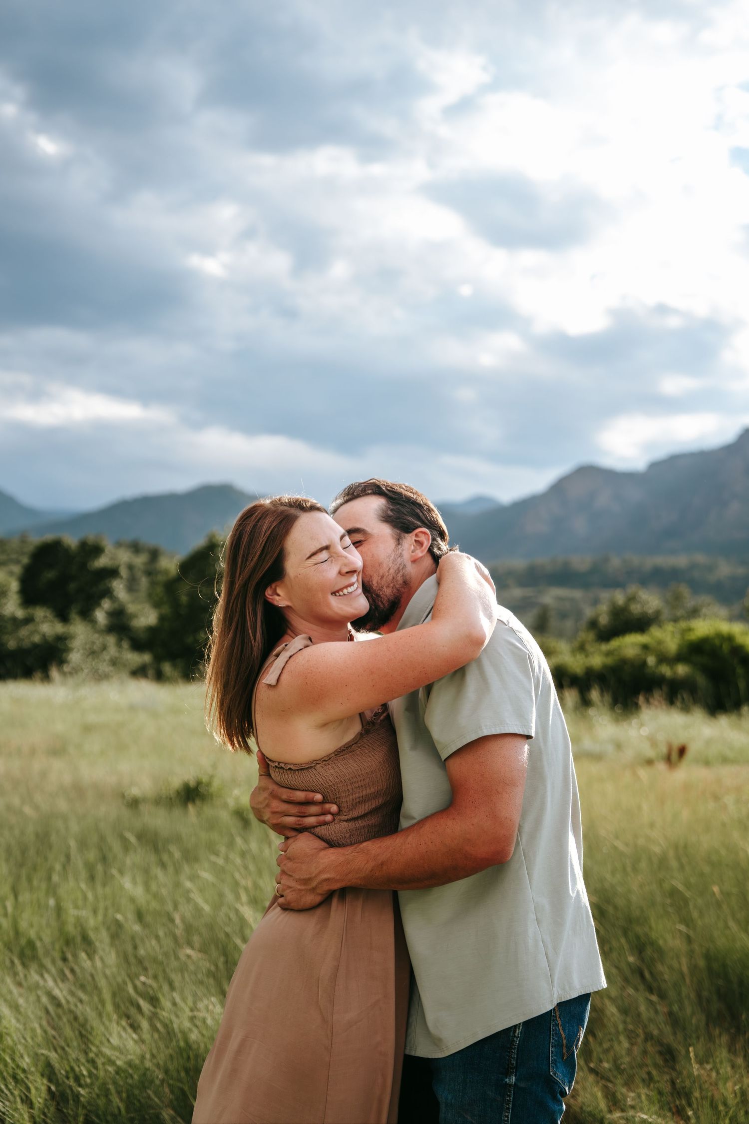A joyful couple embraces in a lush green meadow with mountain views under a cloudy sky.