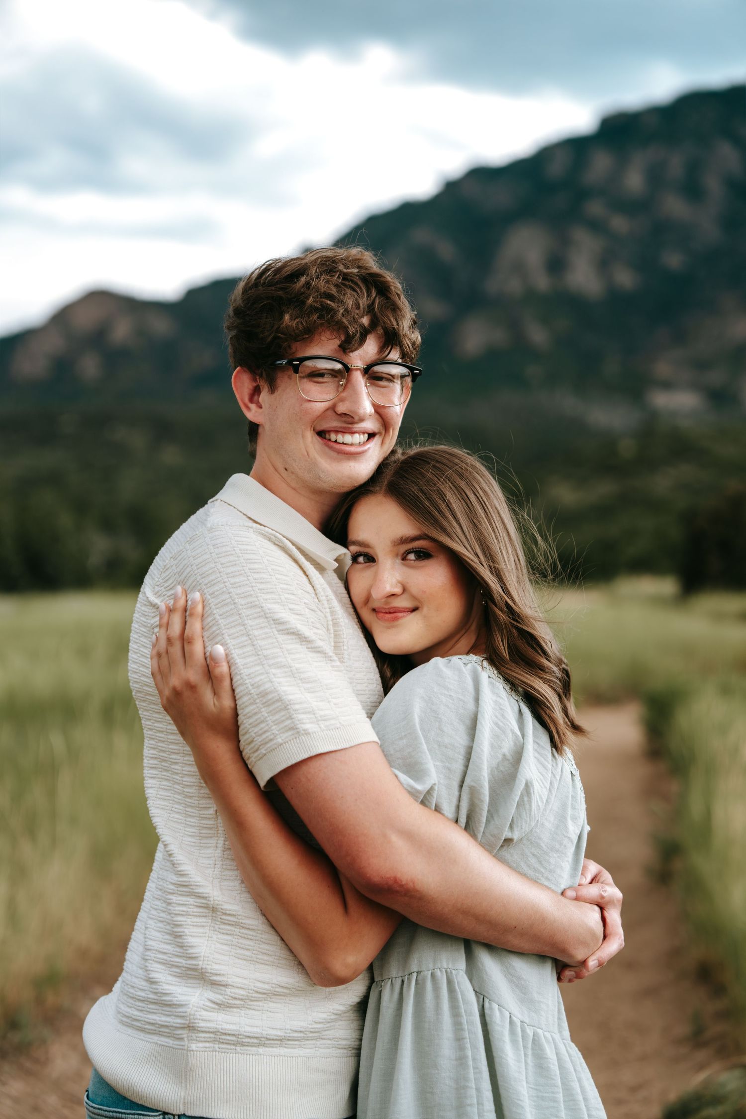 Smiling couple embracing in a green mountain field, man wearing glasses and white polo shirt.