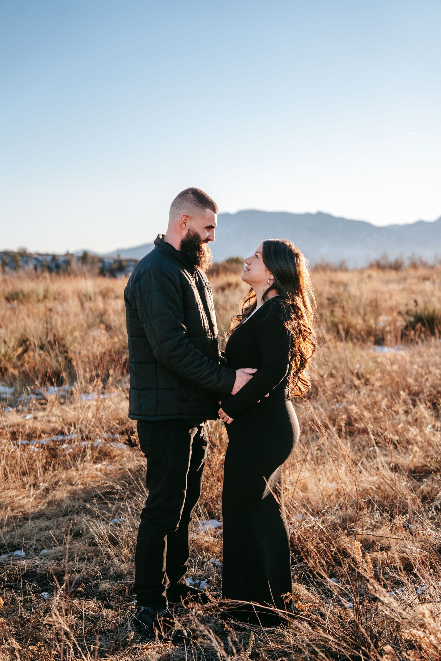 Expecting couple in all black outfits gazing at each other lovingly in golden winter field.