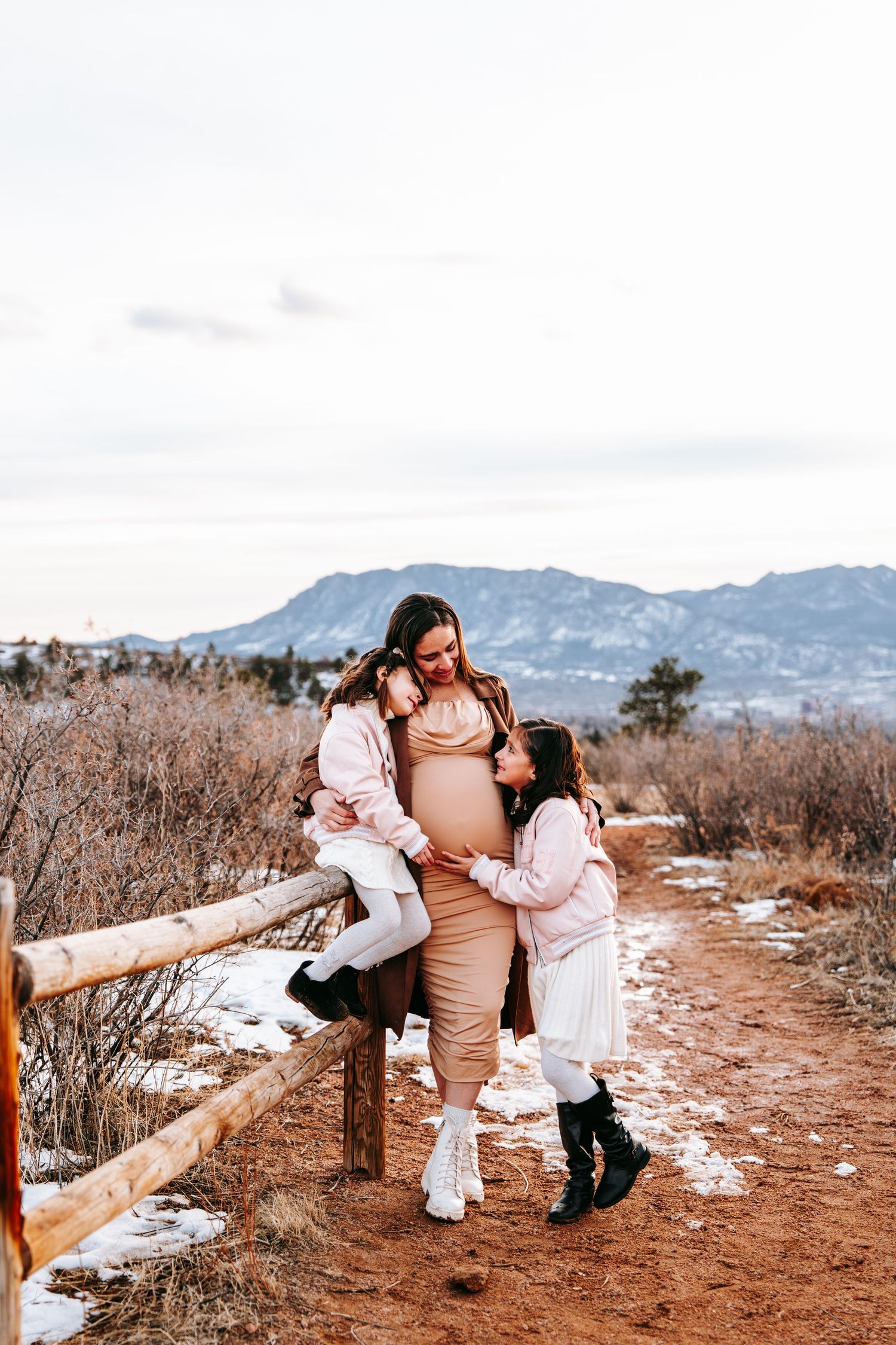 Pregnant mother in beige dress with two daughters near rustic fence, mountains visible in winter landscape background.