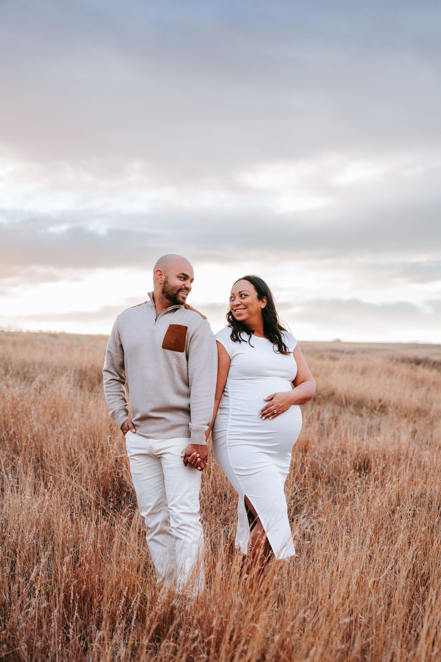 Smiling expecting couple holds hands while walking through golden dry grass field at sunset.