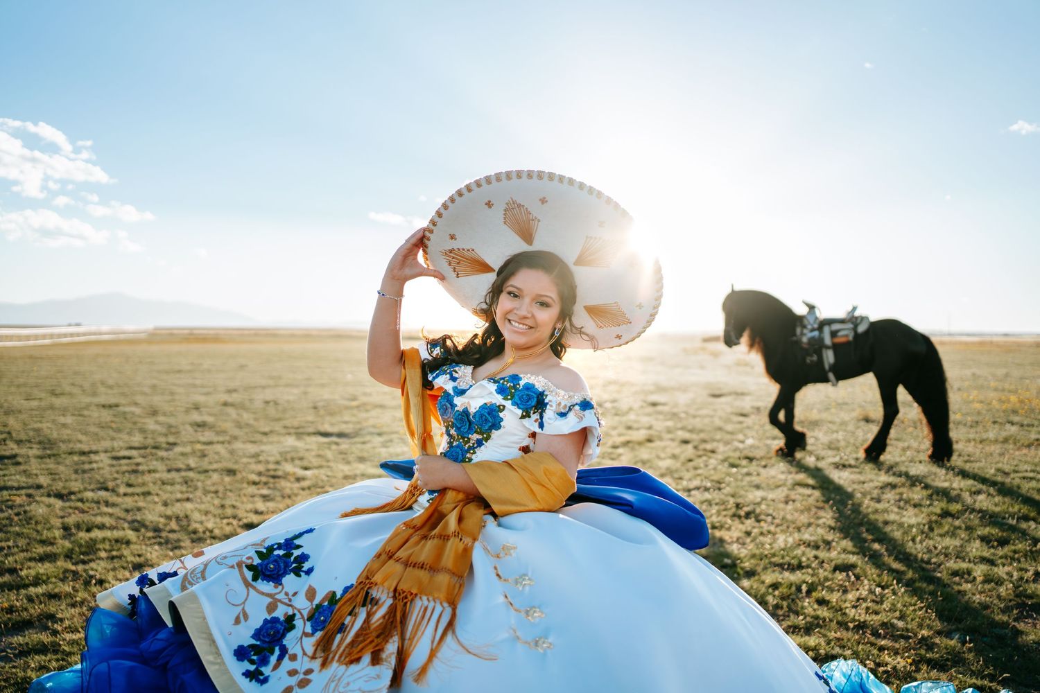 Smiling quinceañera in white blue gold dress holding sombrero in sunny field with black horse.