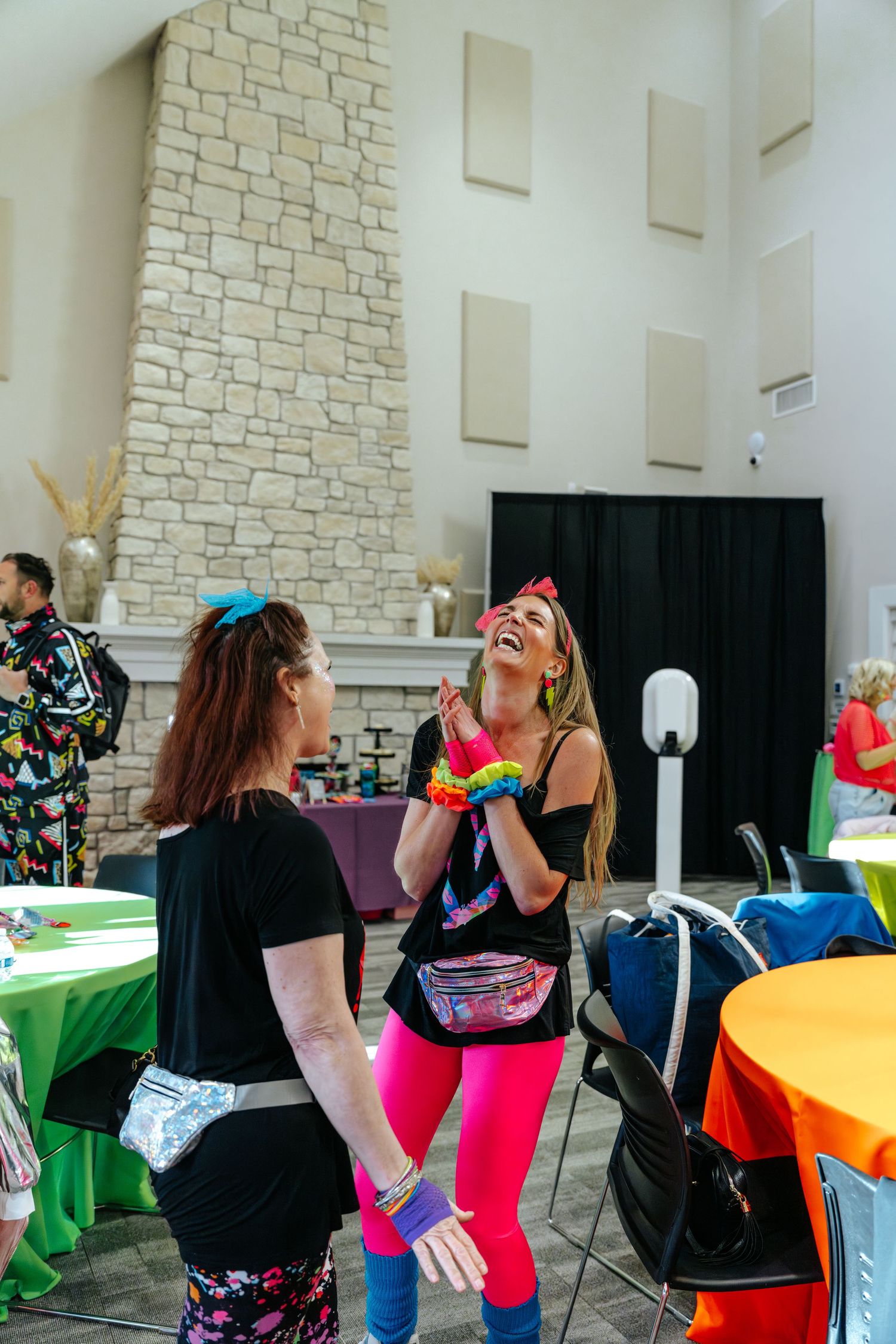 Two women in colorful 80s-themed outfits laugh together at a festive indoor event with decorated tables.