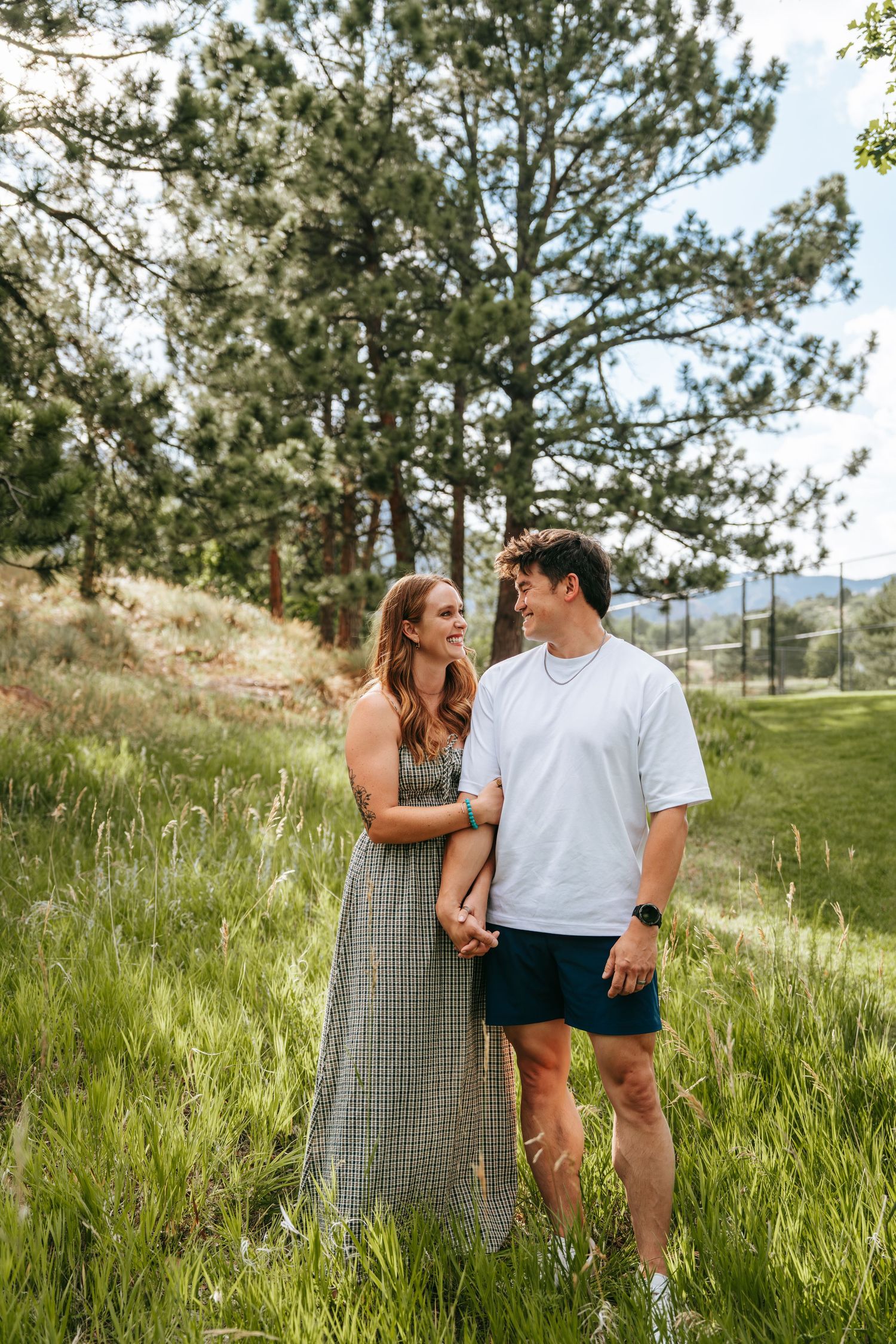 A couple holds hands and smiles at each other in a lush green meadow surrounded by tall pine trees.