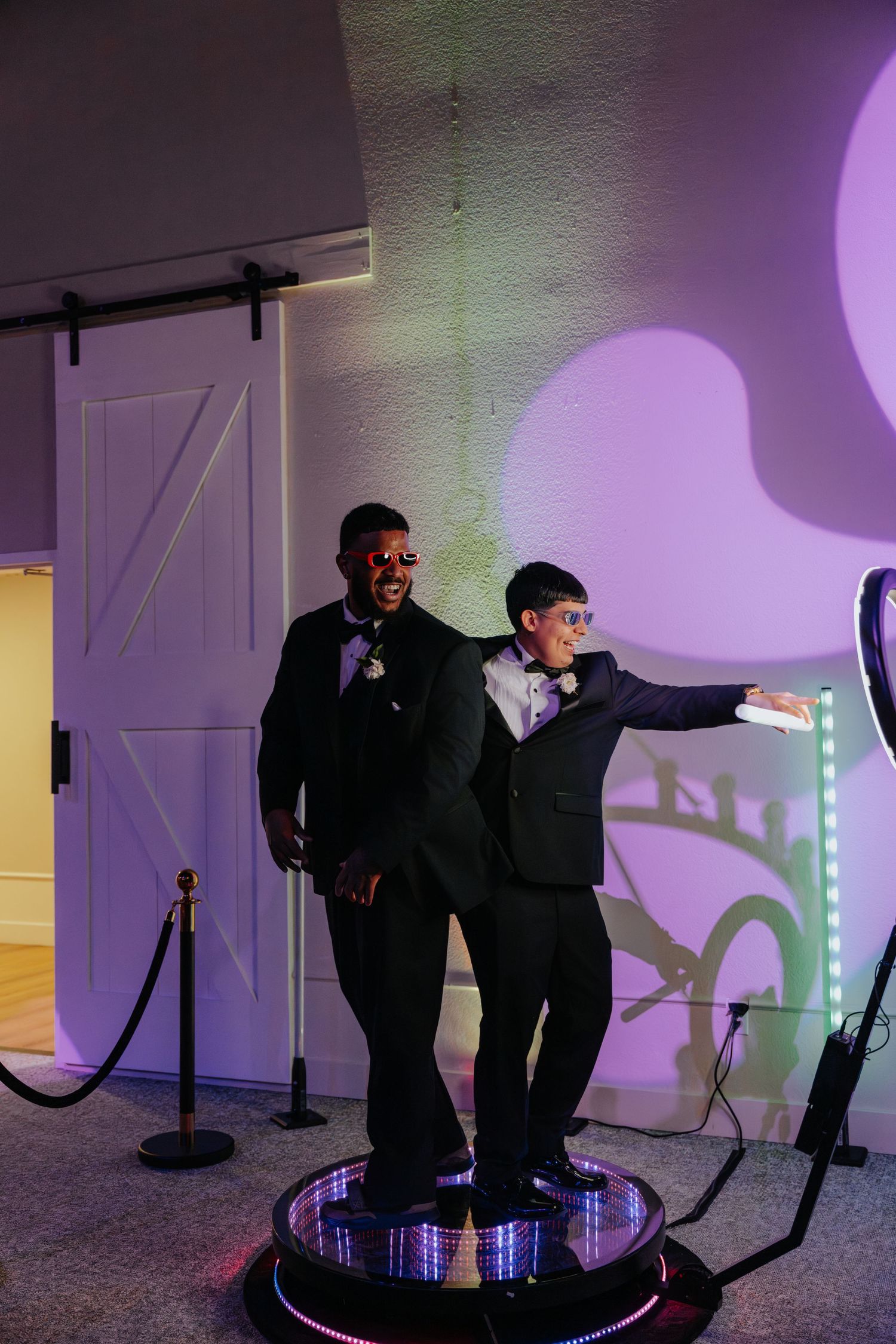 Two men in tuxedos pose on a rotating photo booth platform at a formal event with purple lighting.