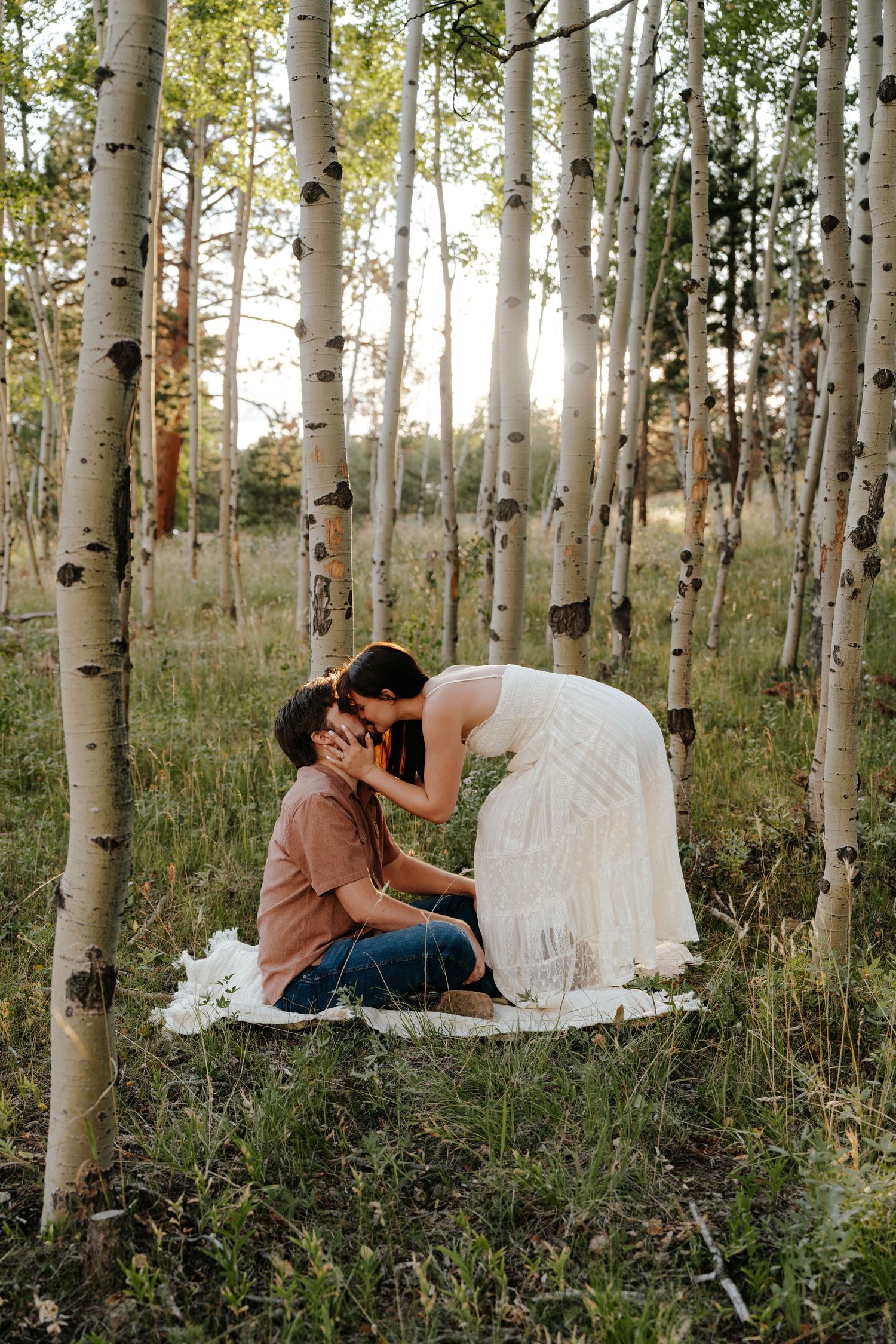 Couple sharing a romantic kiss among white birch trees on a blanket in a sunlit forest meadow.