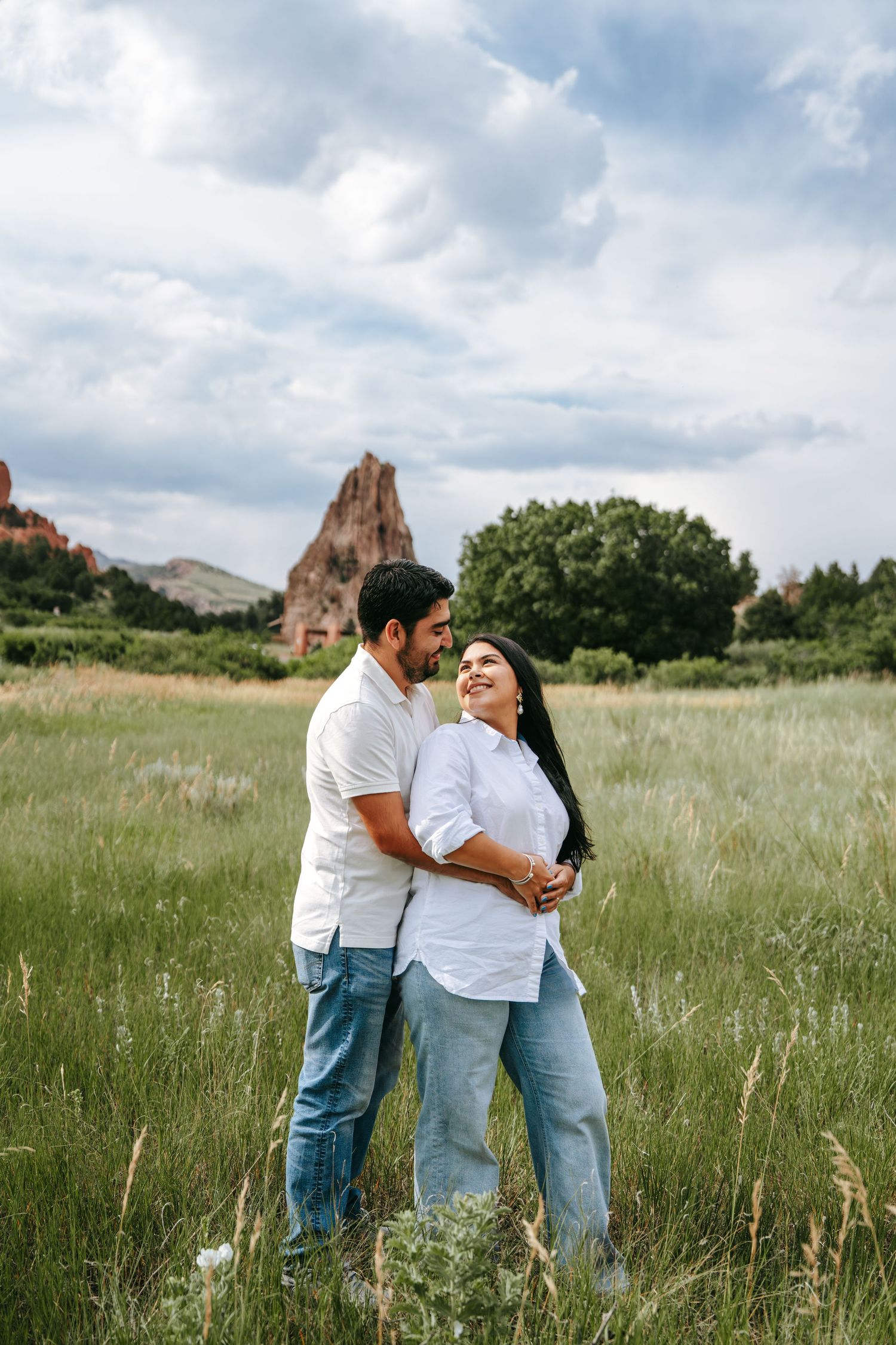 Couple embracing in a green meadow with dramatic red rock formations and cloudy sky in the background.