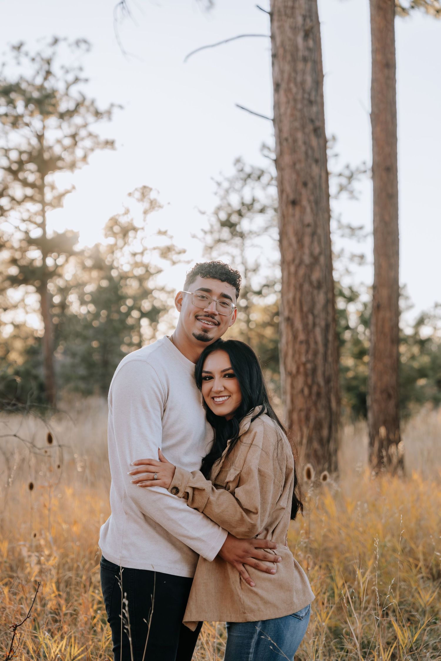 Couple embracing and smiling in a sunlit pine forest field during golden hour engagement photos.
