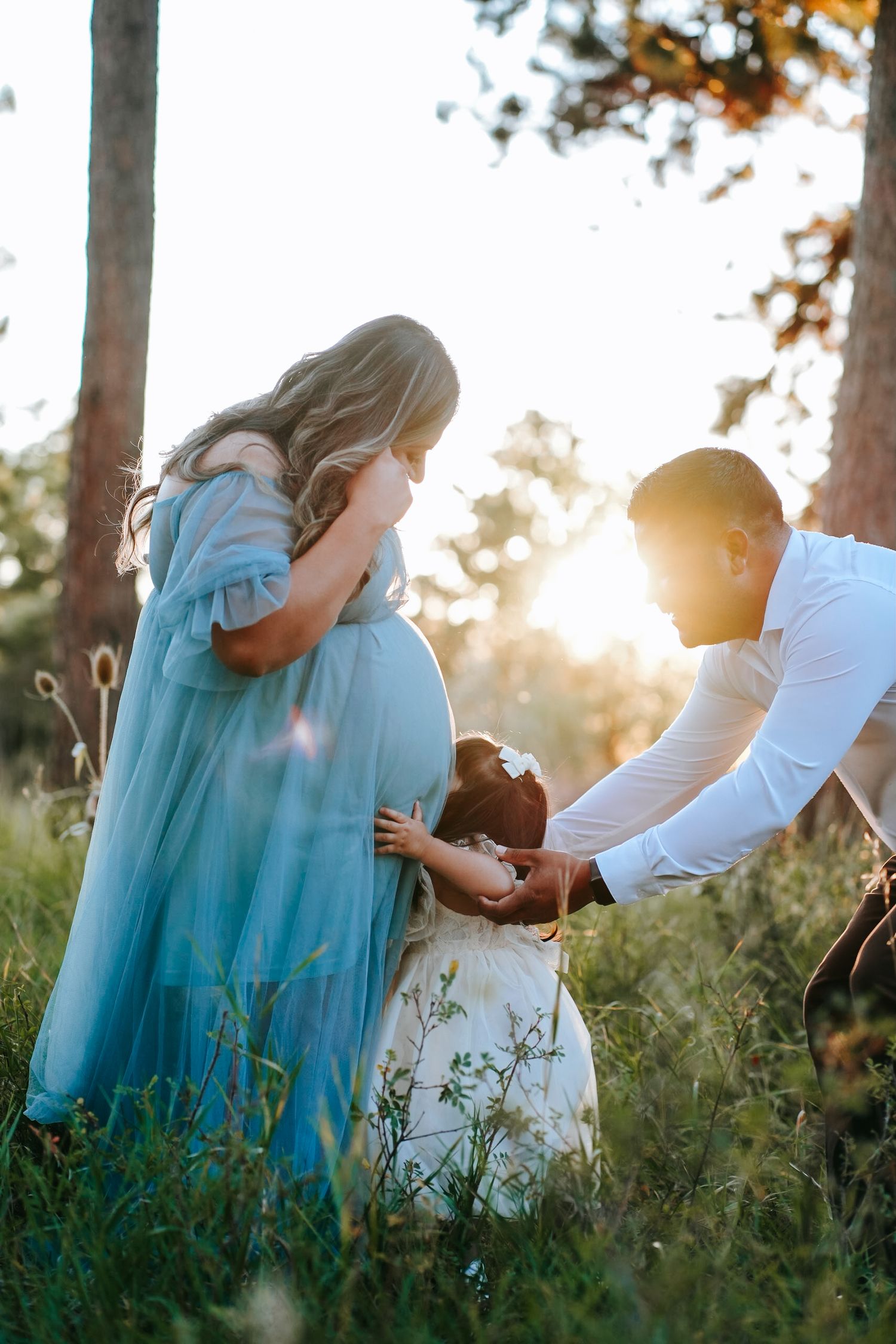 Family maternity photo with pregnant woman in blue gown, man, and child in sunlit forest.