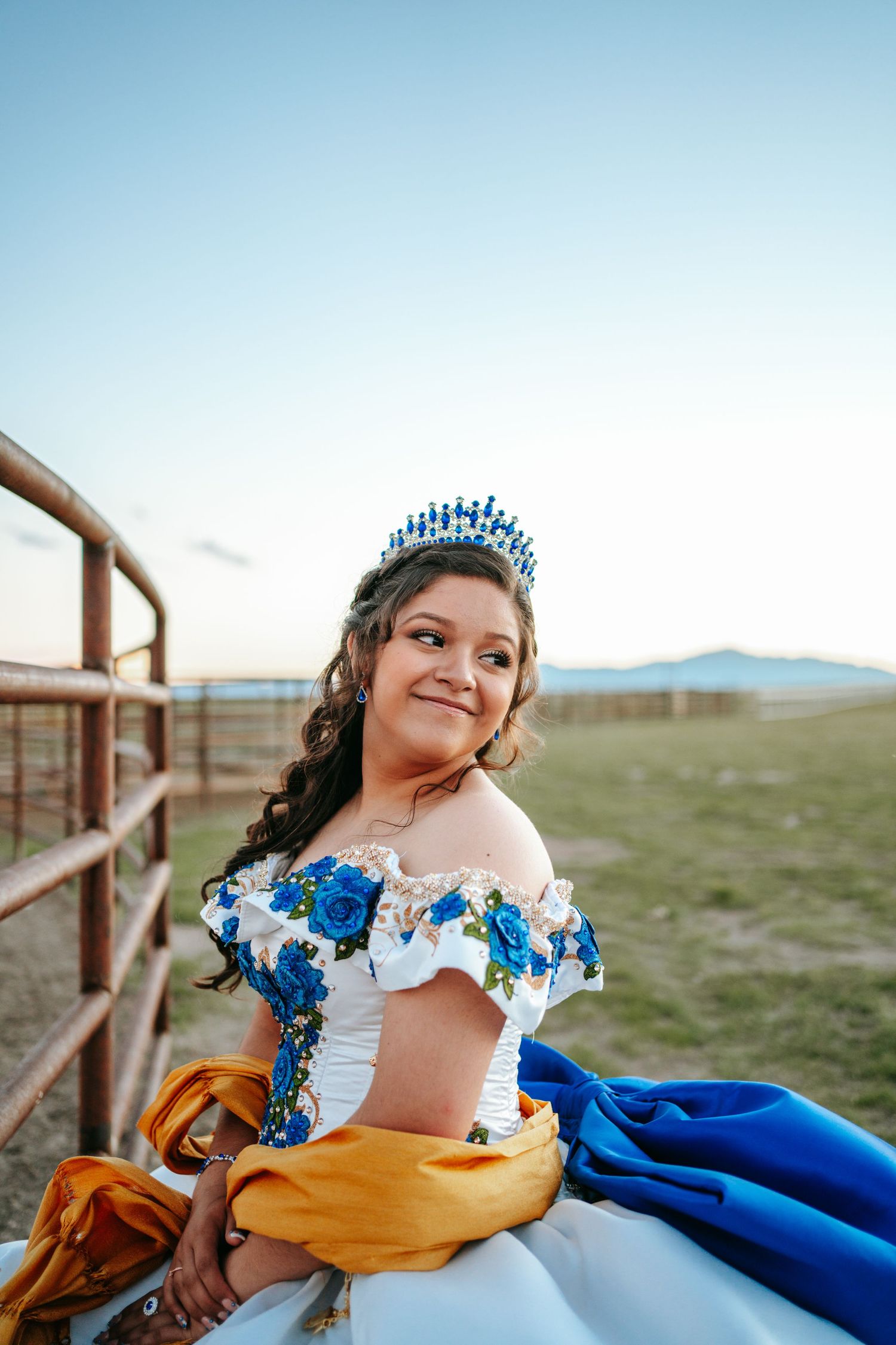 Young woman in quinceañera gown with blue roses and tiara poses by metal fence outdoors.