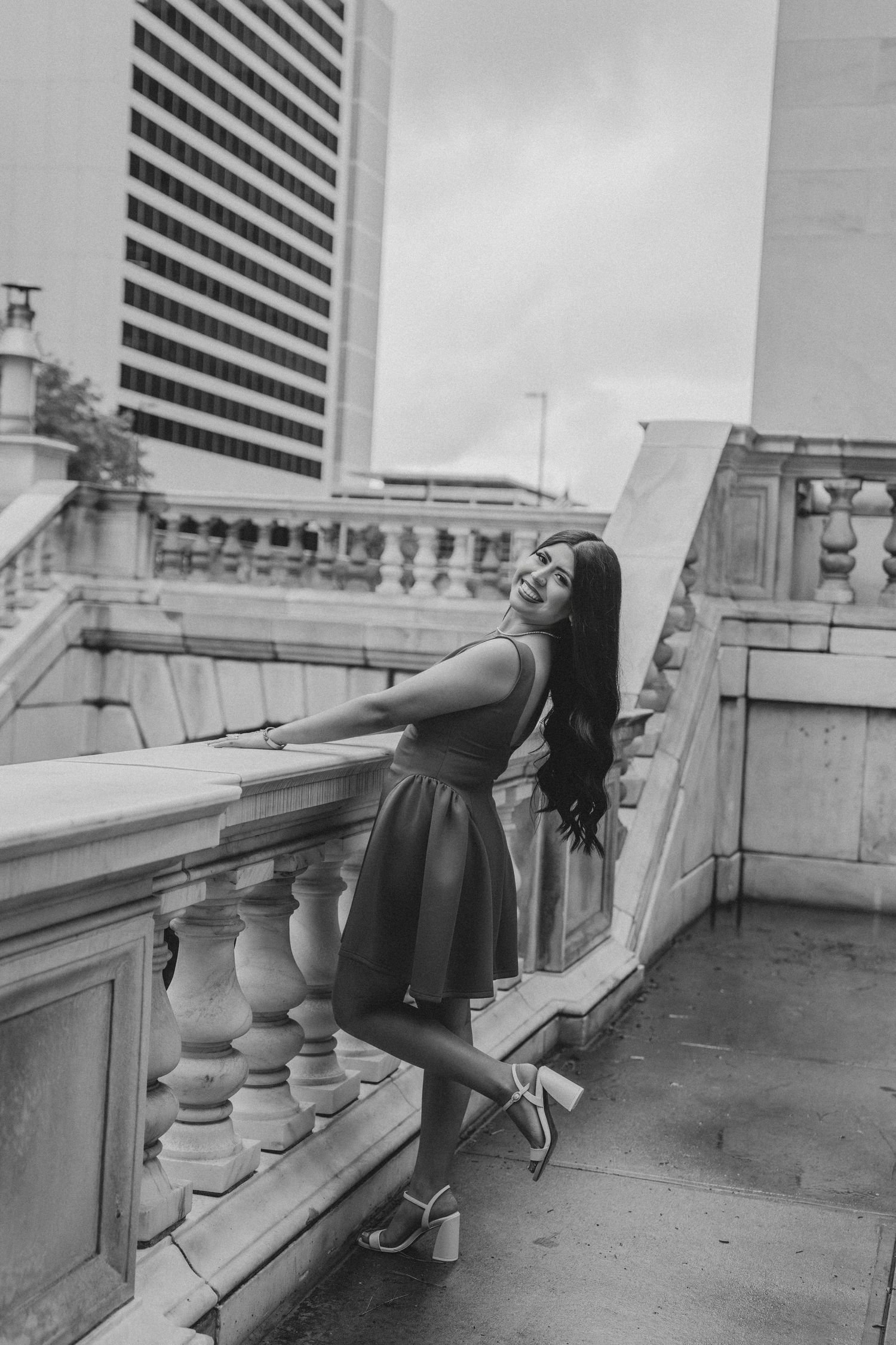 Black and white photo of woman in dress posing on ornate stone balcony with city skyline.