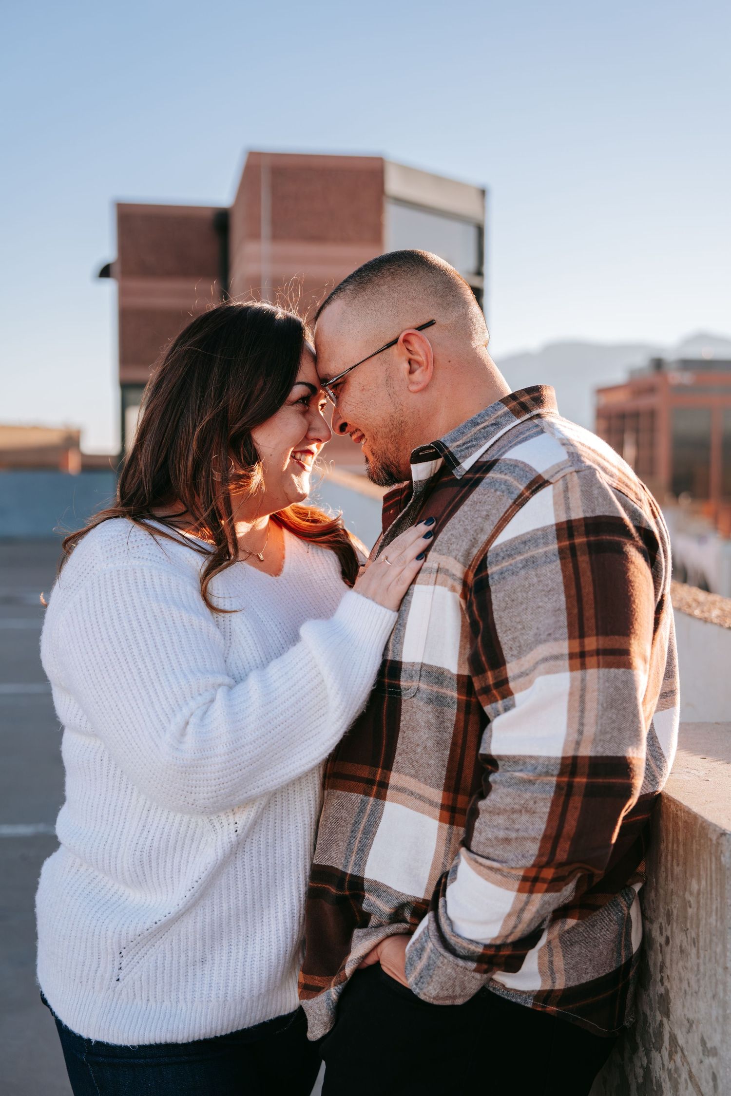 Romantic couple touching foreheads on a rooftop at sunset, woman in white sweater, man in plaid shirt.