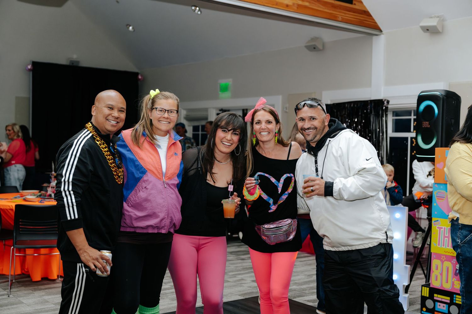 Group of five friends posing together at a colorful 80s themed party event in a decorated venue.
