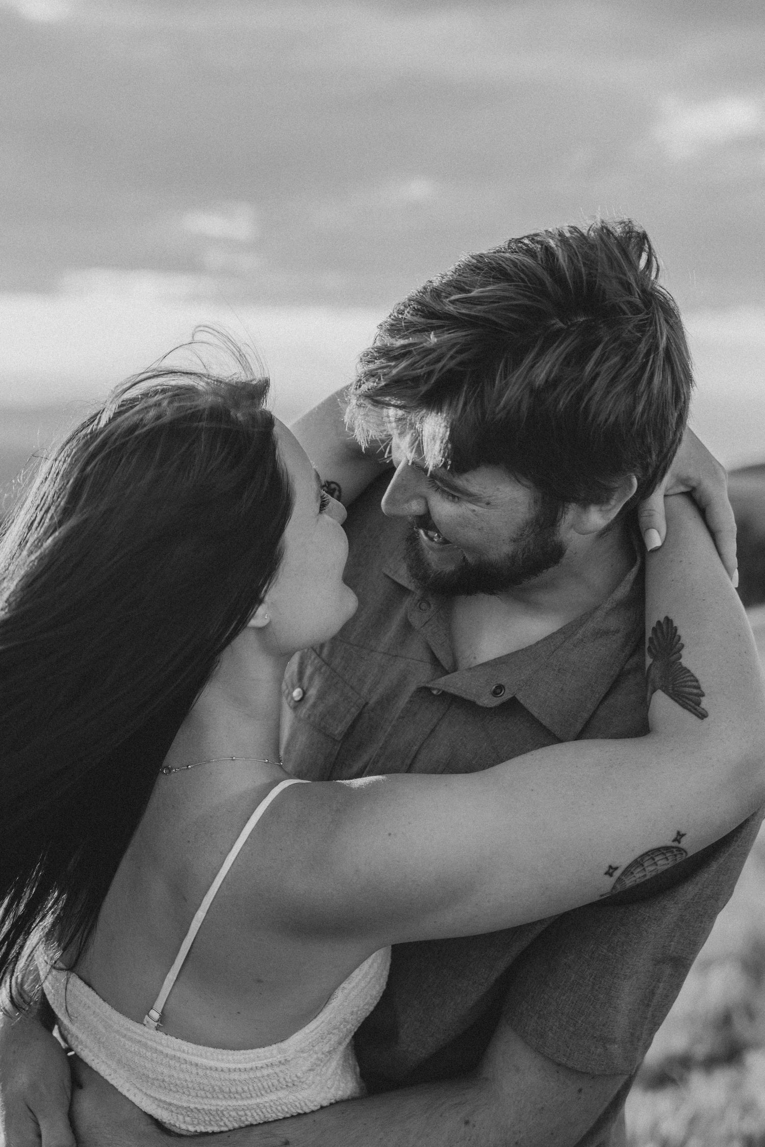 Black and white photo of a romantic couple embracing outdoors, smiling at each other under a cloudy sky.