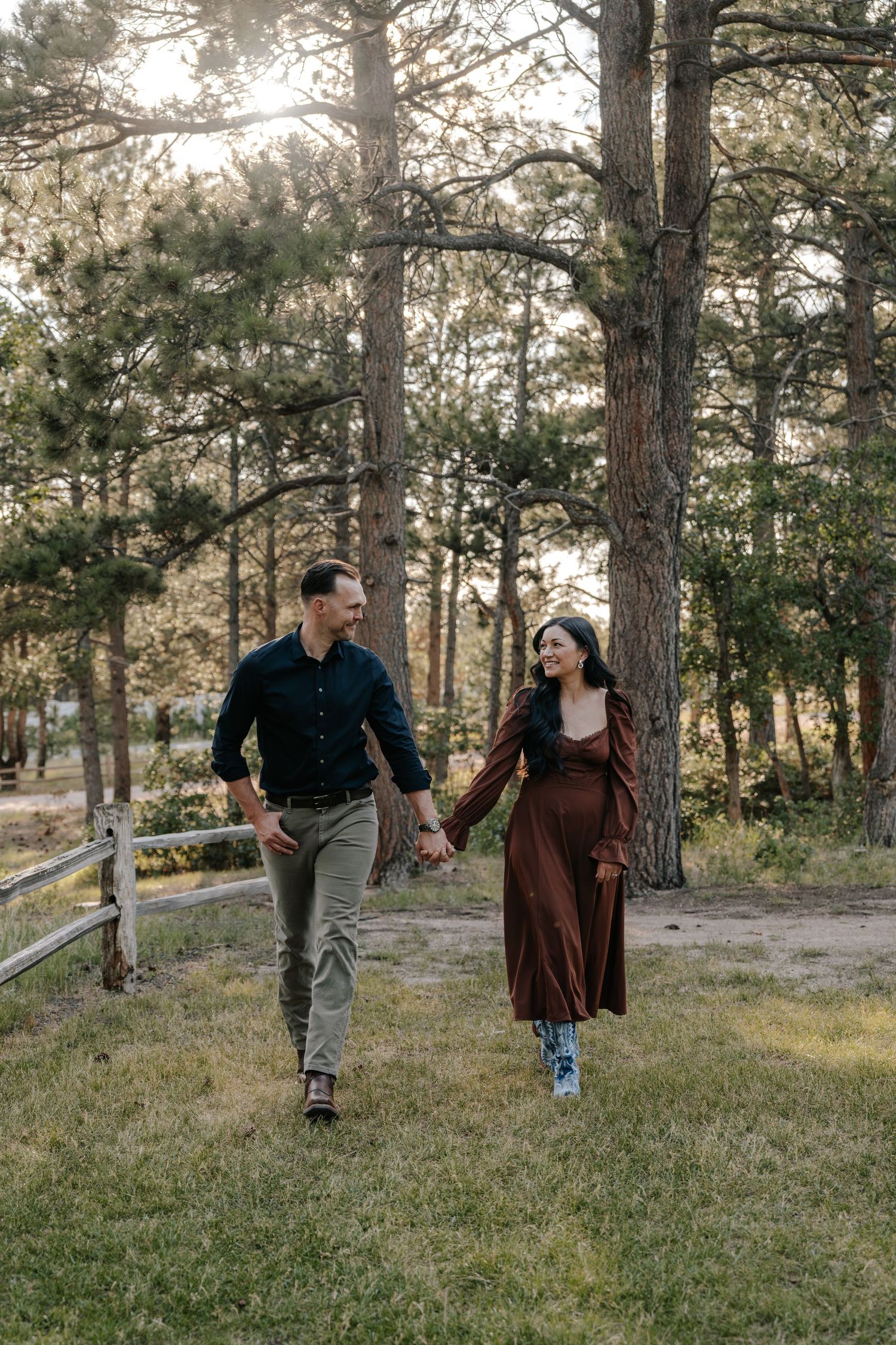A couple holds hands and walks through a sunlit pine forest, the woman in a brown dress and the man in a dark shirt.